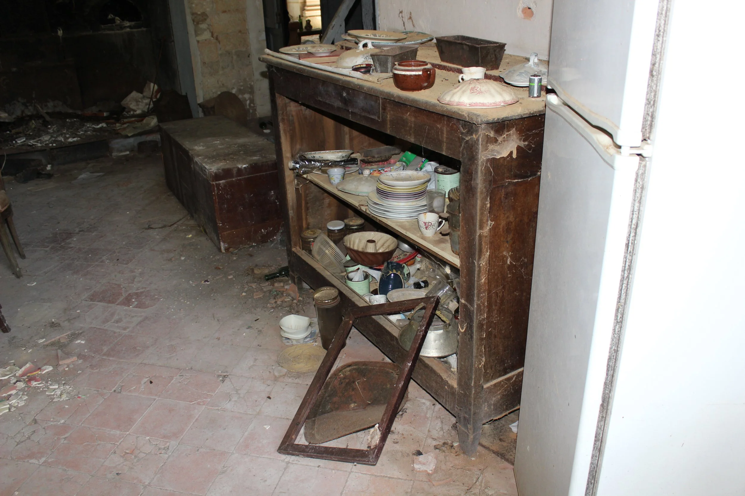 Ein alter, verstaubter Küchenschrank mit zerbrochenem Glas und schmutzigen Geschirr, neben einem weißen Kühlschrank in einem unsauberen Raum.