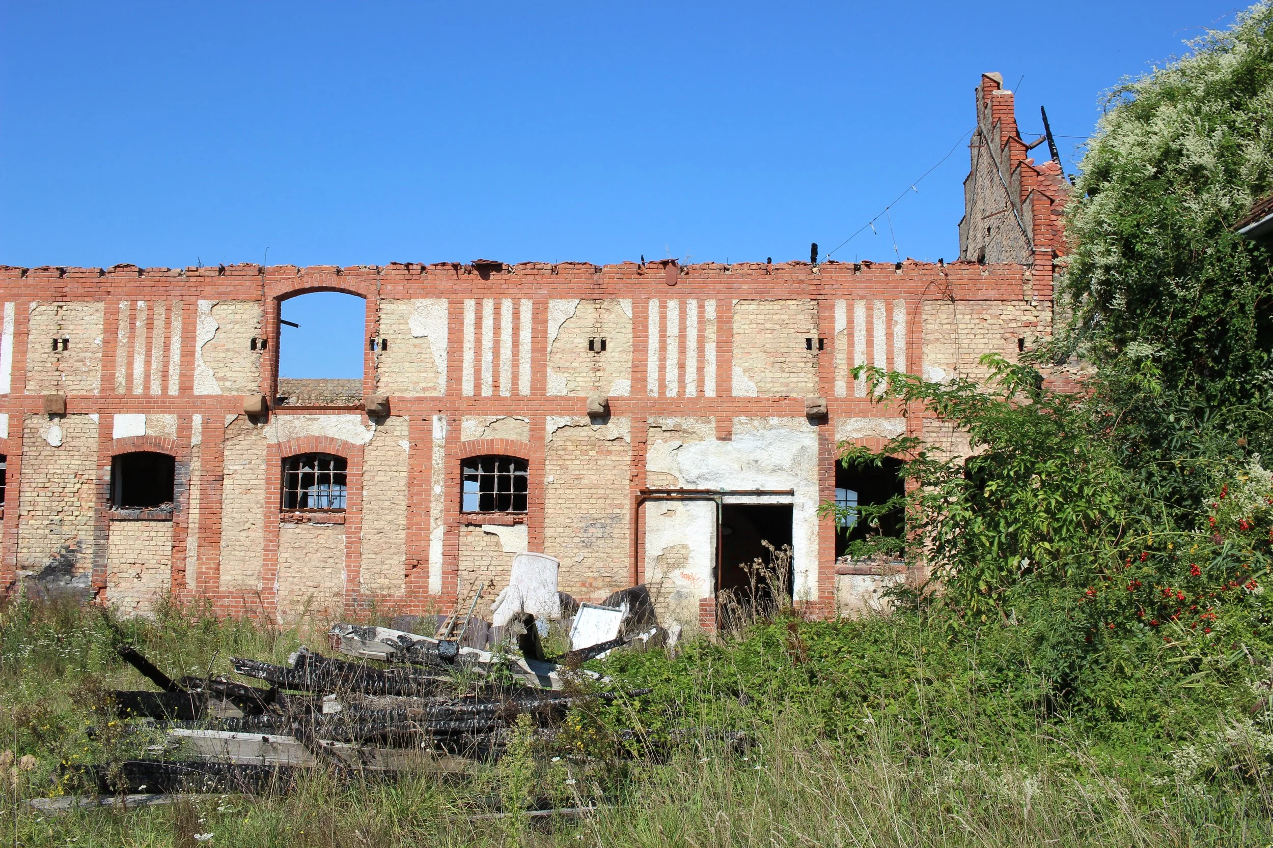 Verlassene, alte Backsteinruine mit offenen Fenstern und überhängenden Wänden. Im Vordergrund Gras und wilde Pflanzen, darunter Müll und Baumaterial. Blaues, wolkenloses Himmel im Hintergrund.