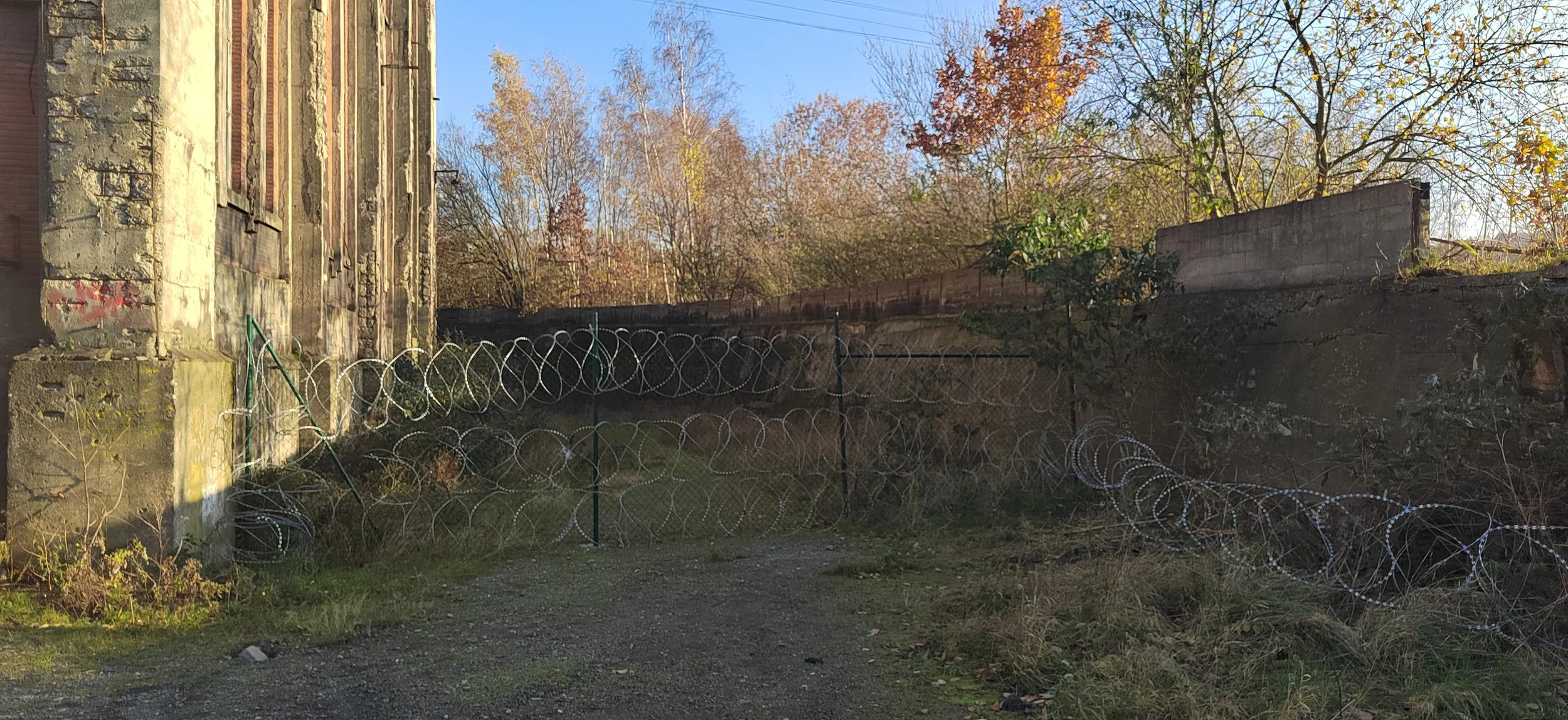 Ein verwitterter, schlossartiger Gebäudeteil auf der linken Seite, umgeben von Stacheldrahtzäunen, mit Bäumen im Hintergrund, bei Tageslicht mit blauem Himmel.
