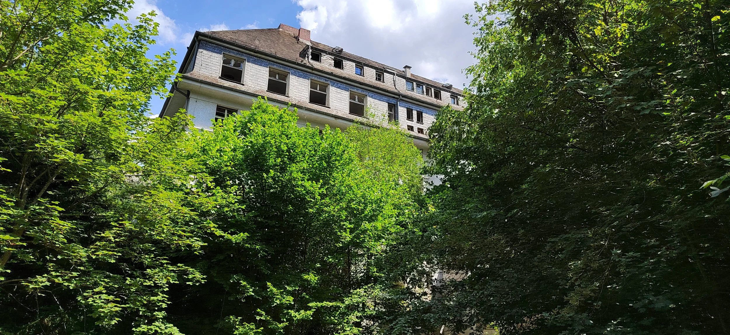 Ein verfallenes mehrstöckiges Gebäude, umgeben von üppigem Grün mit vielen Bäumen, unter einem bewölkten Himmel.