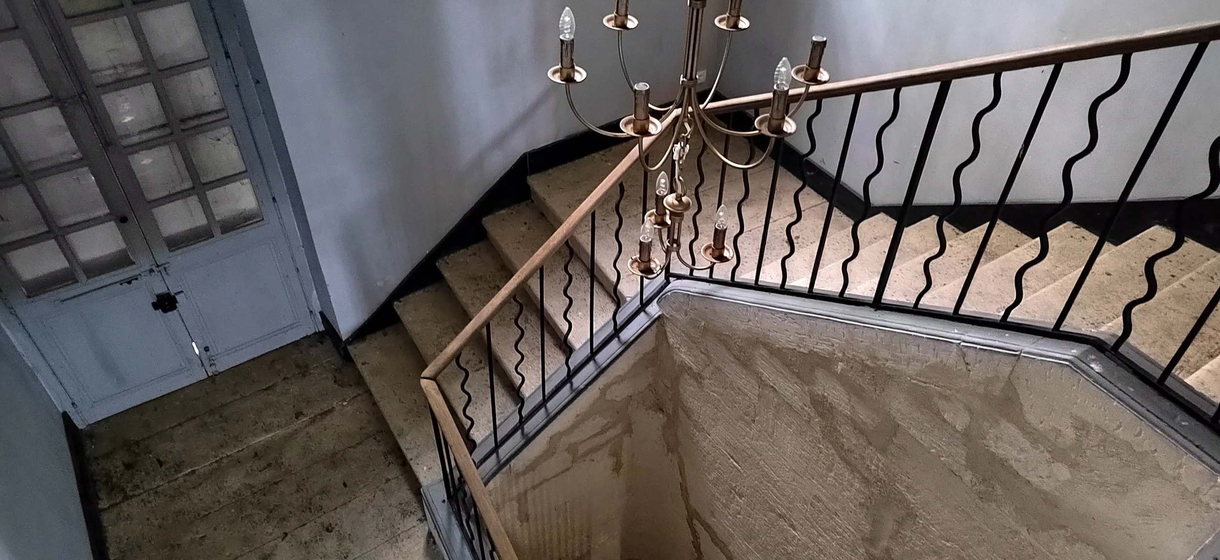 Blick auf ein geschwungenes Treppenhaus mit einer hölzernen Handlauf und einer dekorativen Kronleuchter mit Glühbirnen. Im Hintergrund ist eine weiße Tür mit Glasfenstern.