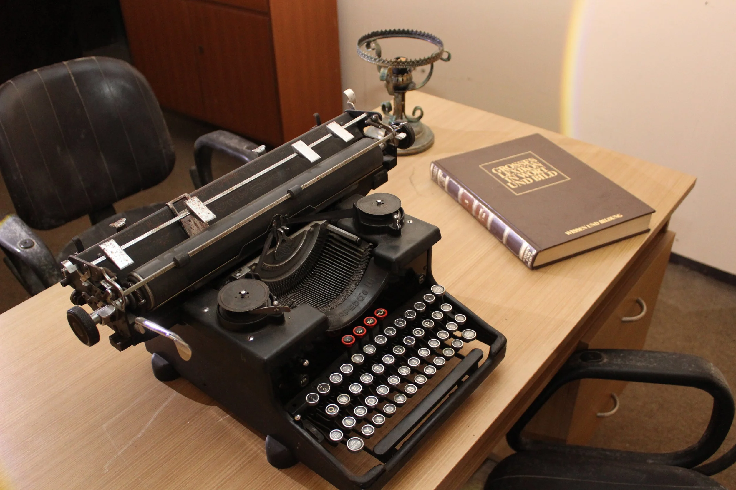 Antike Schreibmaschine auf einem Holztisch, daneben ein Buch und ein Kerzenhalter, im Hintergrund ein Stuhl.