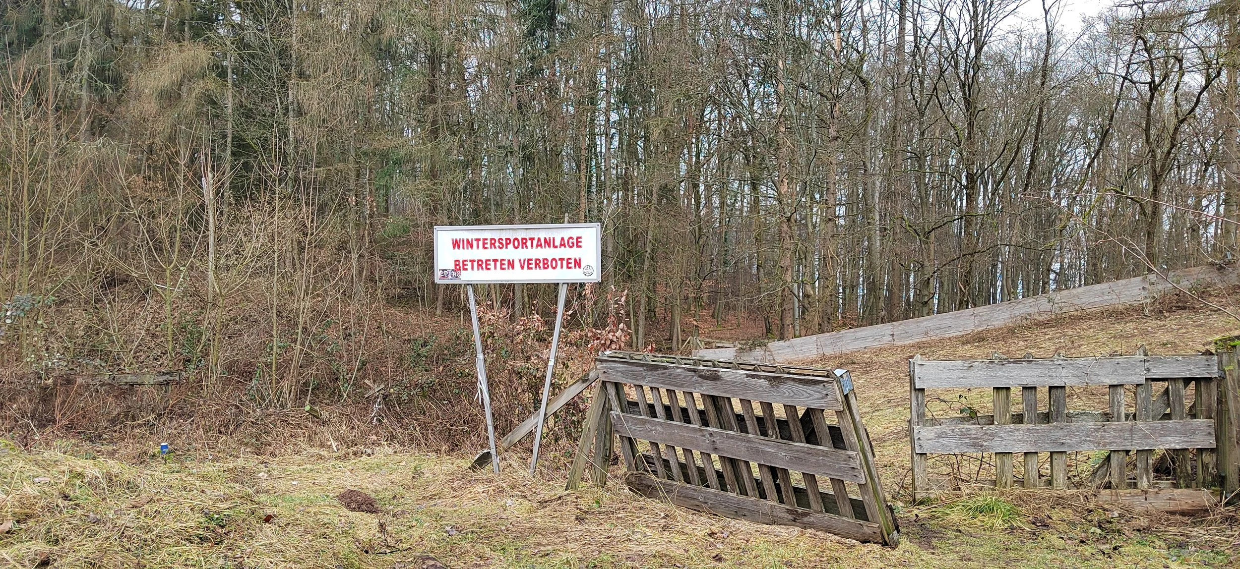 Ein Schild mit roter Schrift auf weißem Hintergrund, das sagt 'WINTERSPORTANLAGE BETRETEN VERBOTEN', steht im Wald neben einer beschädigten Holztür. Der Wald ist kahl, weil es Winter ist.