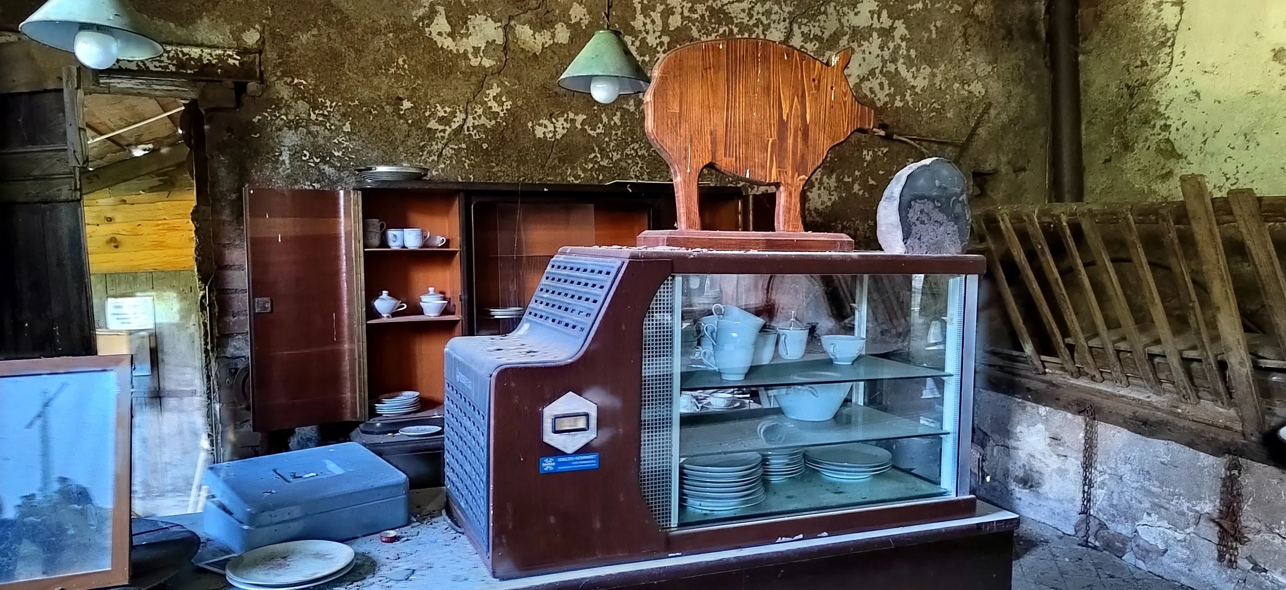 Alte, verstaubte Kücheneinrichtung mit Geschirr, einem Holzstuhl, einem Glaskasten mit Geschirr, und einem Wandregal, in einem rustikalen Raum, vermutlich in einem Lager oder Schuppen.