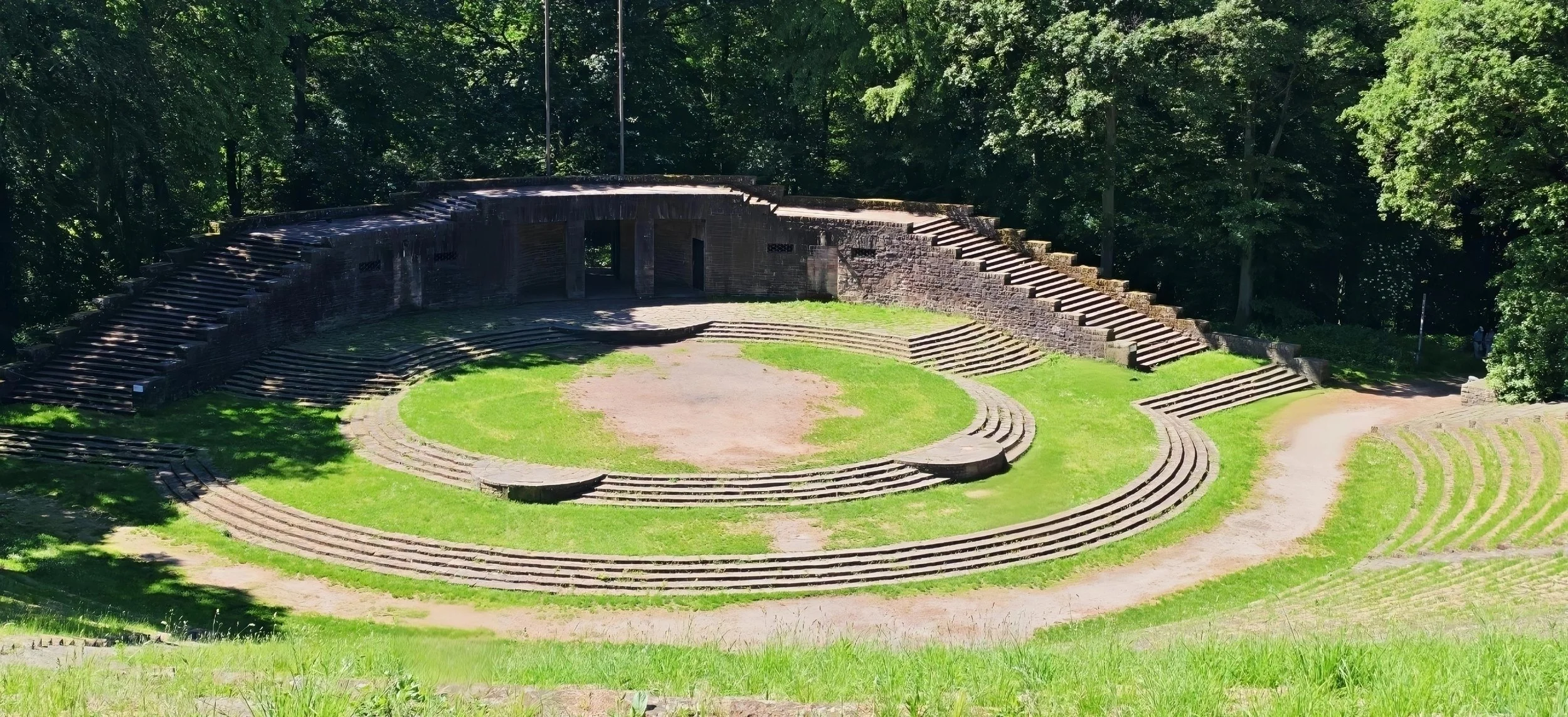 Eine antike römische Arena aus Stein, umgeben von grüner Wiese und Bäumen, im Freien