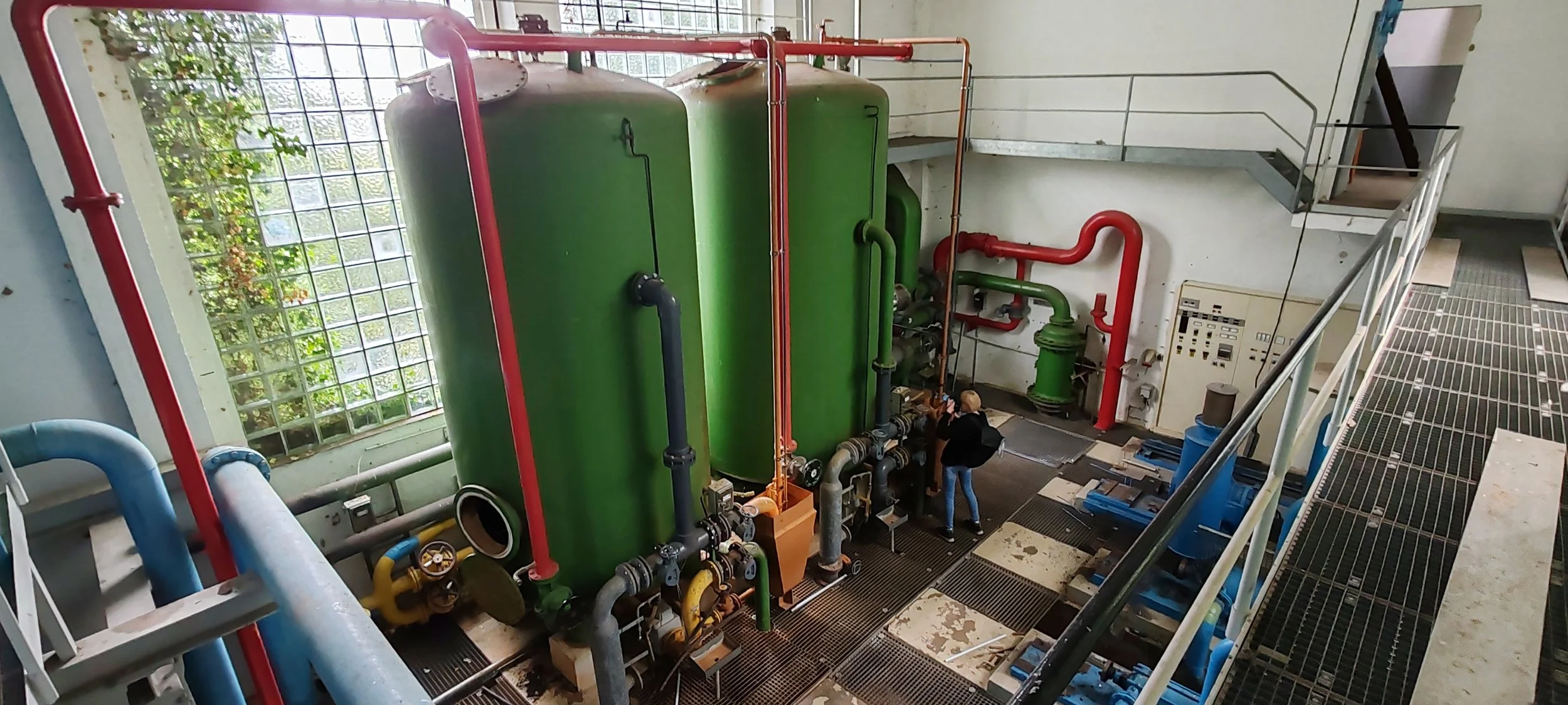 Blick auf Wasseraufbereitungsanlage mit grünen Behältern, roten und grünen Rohren, Bedienfeld, Metallgeländer und Person, die die Anlage inspiziert.