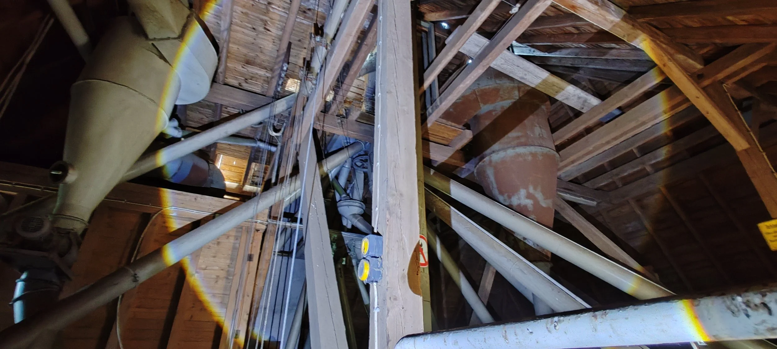 Innenansicht eines Dachbodens unter einem Dach mit sichtbaren Holzstämmen und Rohrleitungen, sowie elektrische Verkabelung.