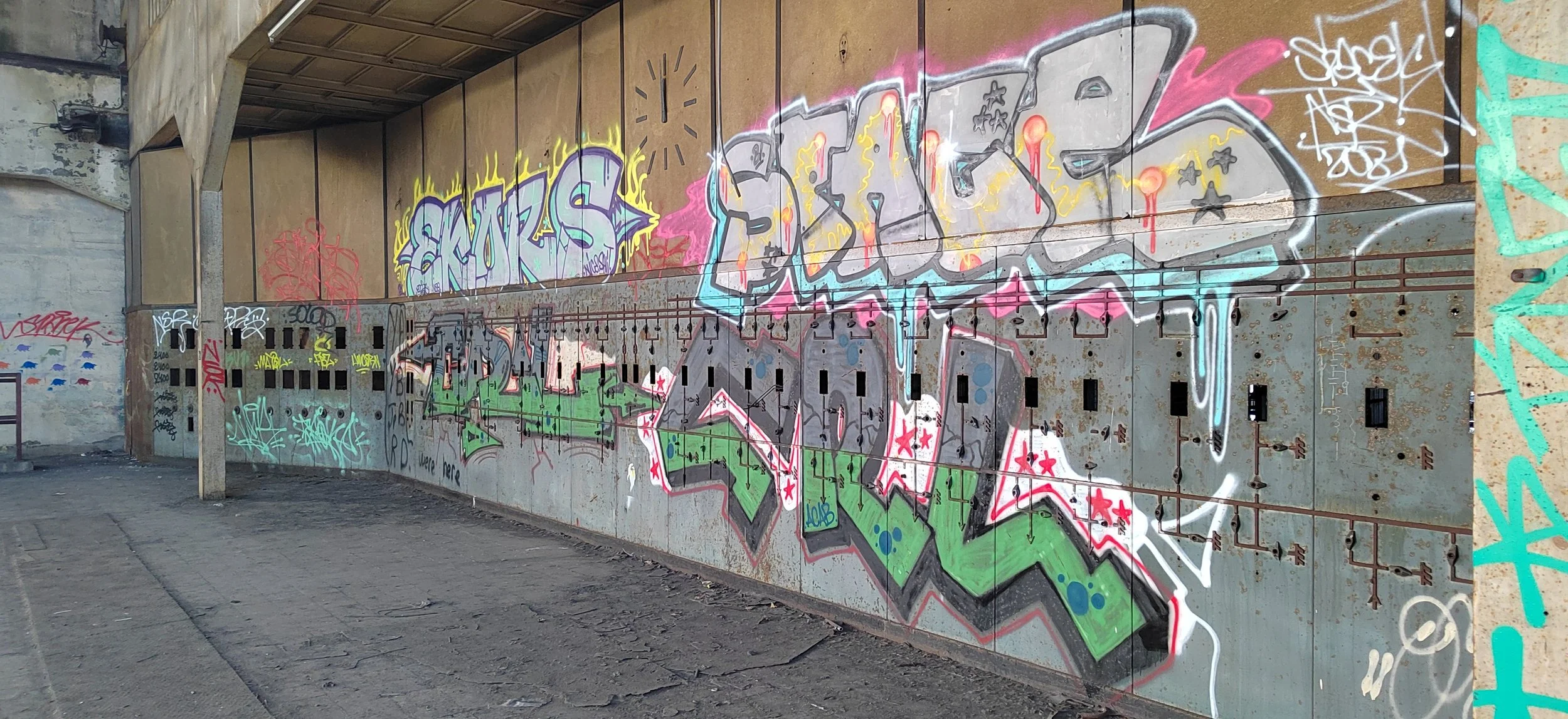 Graffiti an einer Metallwand in einem urbanen Umfeld, bestehend aus mehreren farbigen Tags und Schriftzügen.