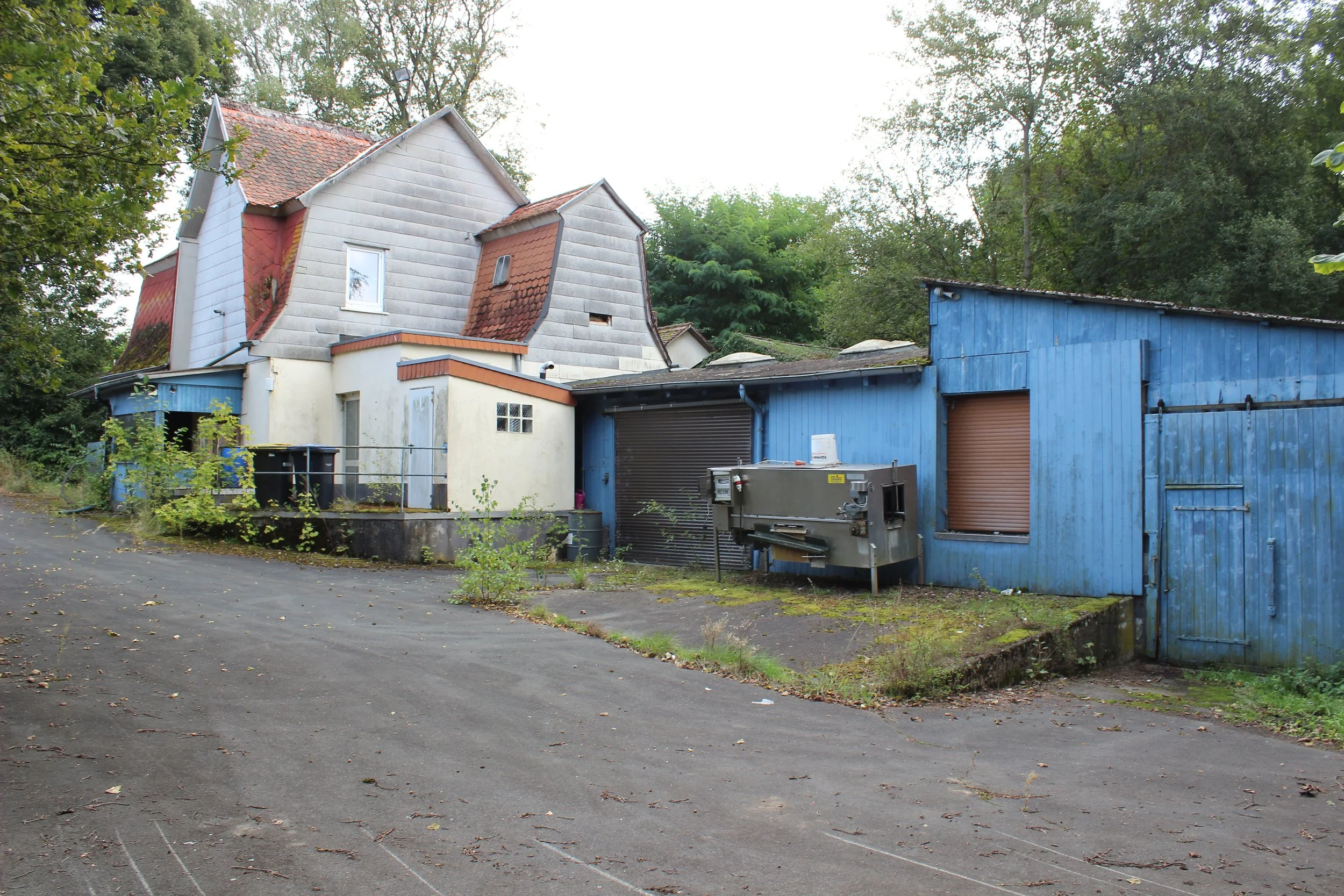 Ein Gebäude mit blauer Holzfassade und einem weißen Haus im Hintergrund, umgeben von Bäumen, auf einer gepflasterten Fläche mit kleinen Pflanzen und Mülltonnen.