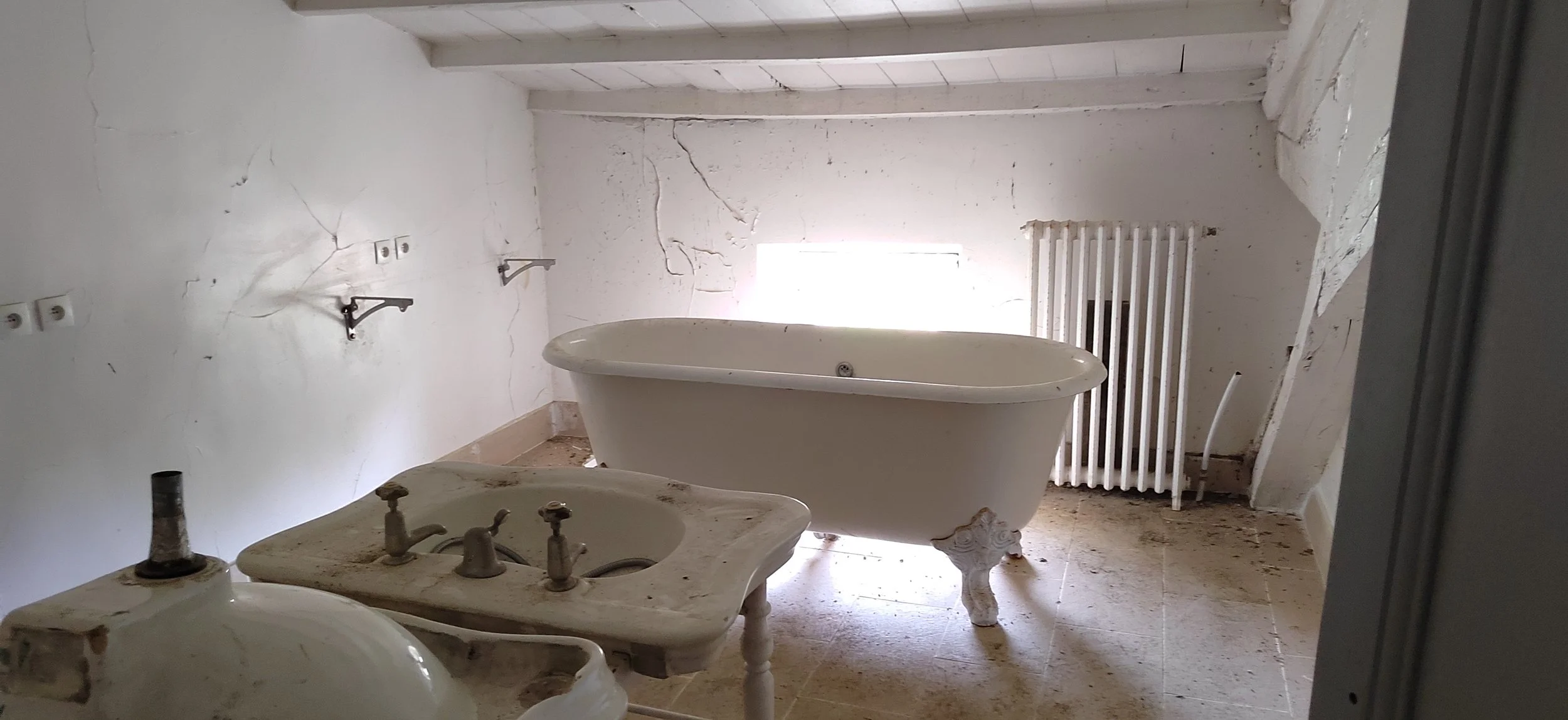 Ein verlassener Badezimmerraum mit einer alten Wanne, einer Spüle und einem Heizkörper, alle in einem schlechten Zustand, mit Rissen an den Wänden und Staub auf den Oberflächen.