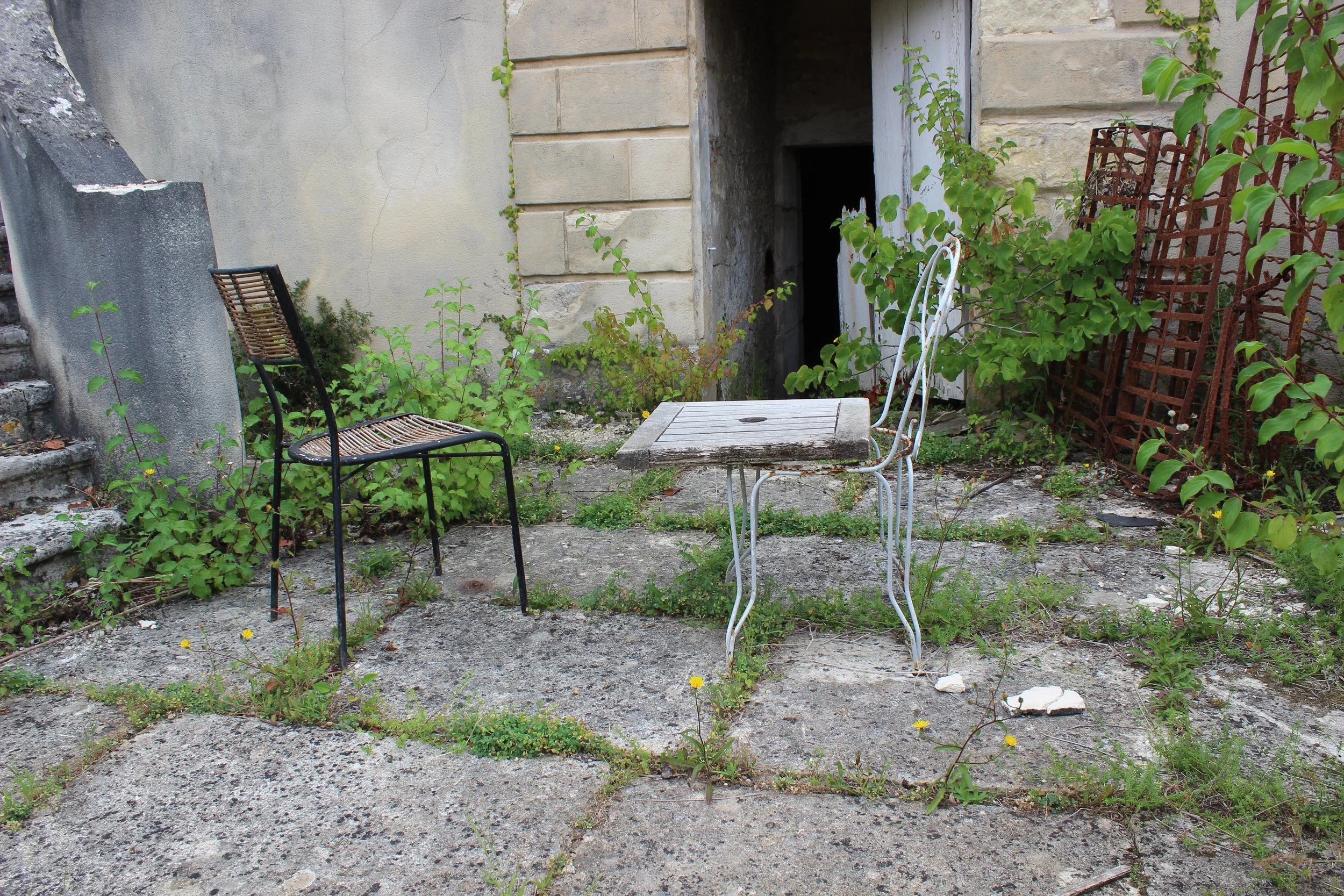 Ein verlassener Hinterhof mit zwei Stühlen, einem kleinen Tisch, Pflanzen und einem dunklen Zugang zu einem Keller oder Kellerraum.