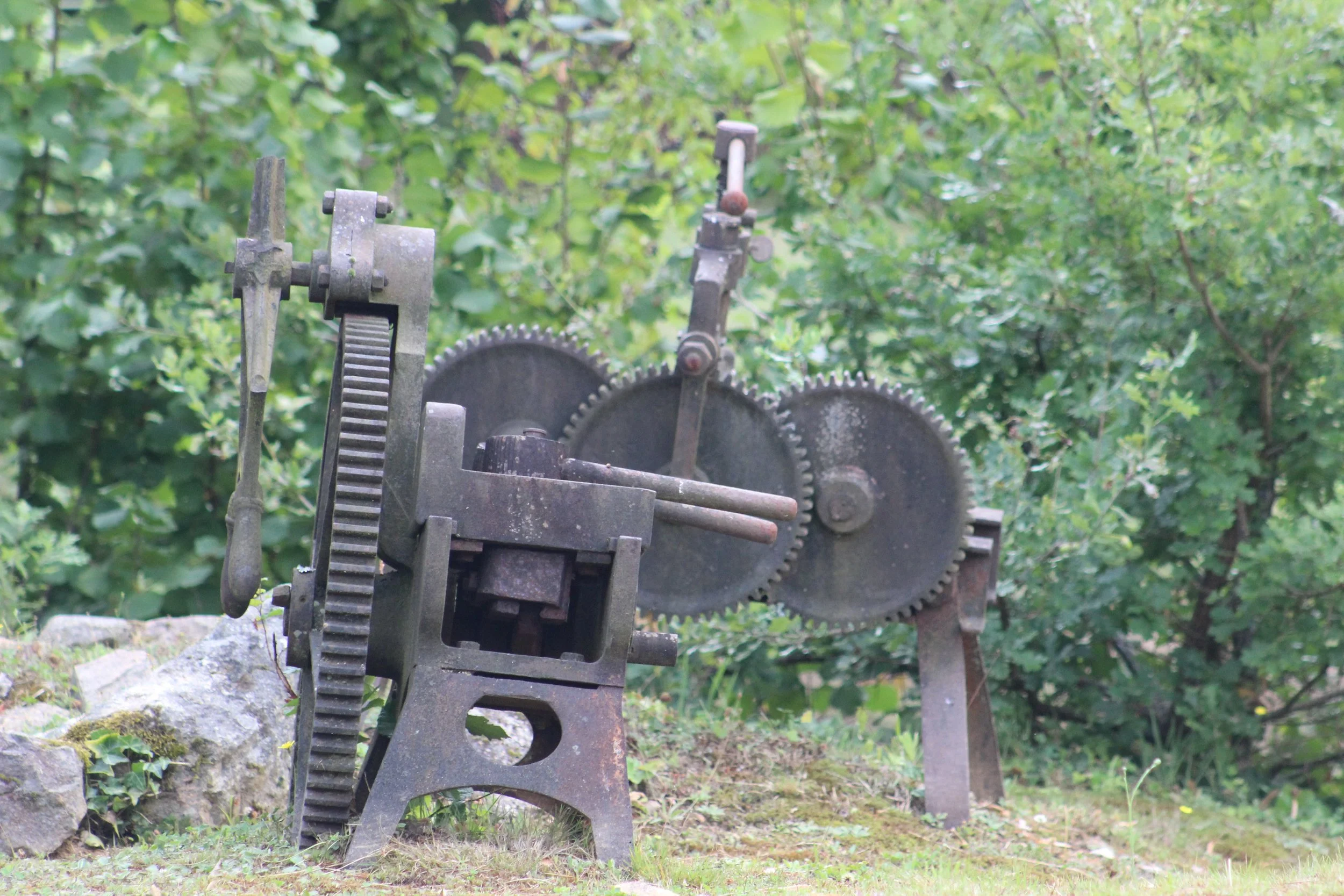 Alte, verrostete mechanische Maschine mit Zahnrädern im Freien, umgeben von grünen Büschen.