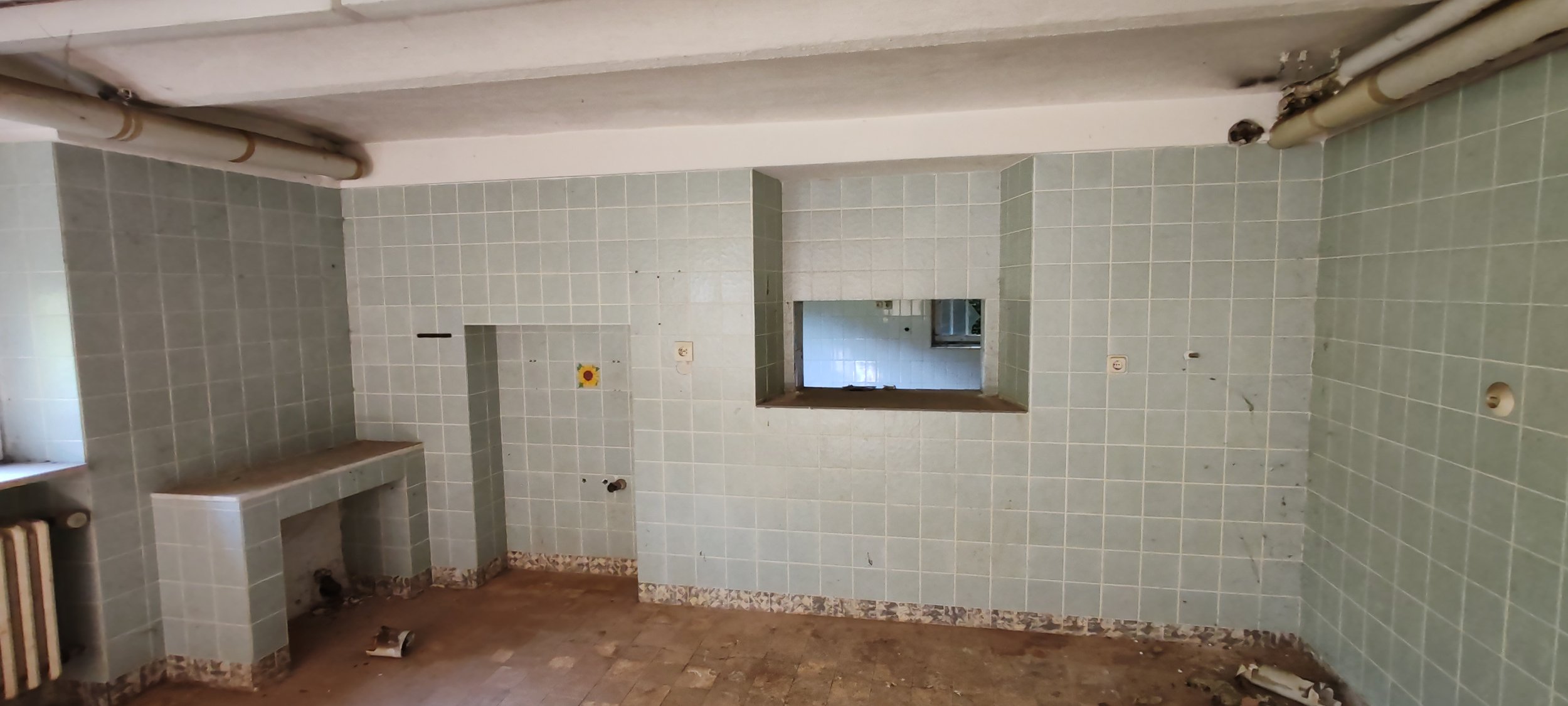 Ein leerer, renovierungsbedürftiger Küchenraum mit grünen Fliesen an den Wänden, einem kleinen Fenster, und einer offenen Küchenöffnung. Der Boden ist uneben und es liegen Baumaterialreste herum.