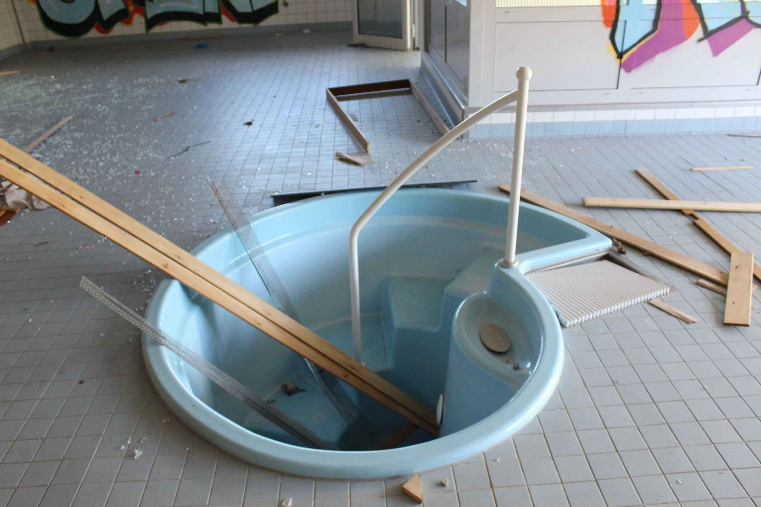 Ein beschädtes, leerstehendes Schwimmbecken mit Holzstücken und Scherben auf dem Boden, in einer vorläufigen Kontrolle