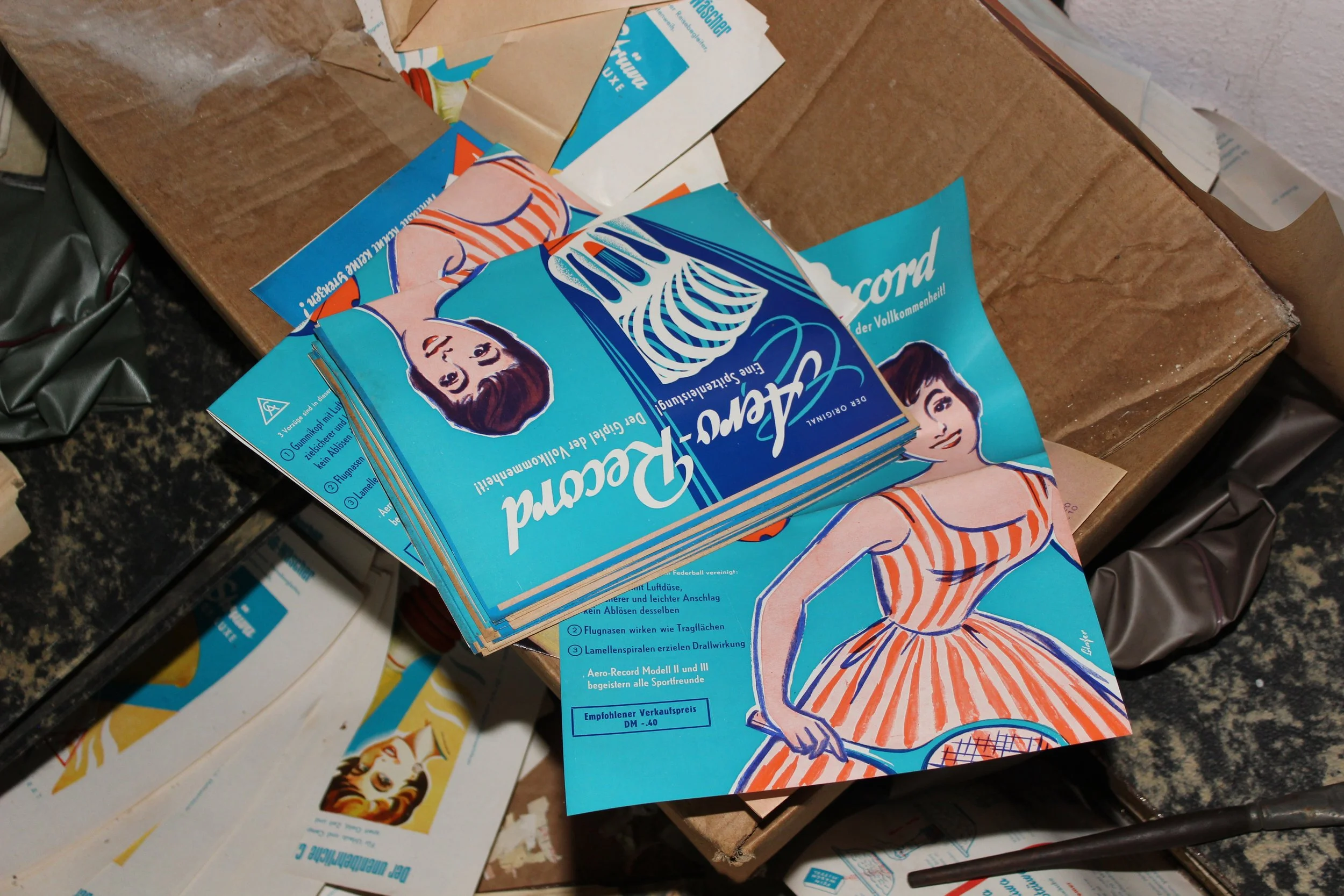 Alte, aufgeblasene Werbe- oder Verpackungsflyer mit einer Illustration einer jungen Frau in einem orange-weiß gestreiften Kleid, die einen Tennisschläger hält, liegen auf einem Tisch.