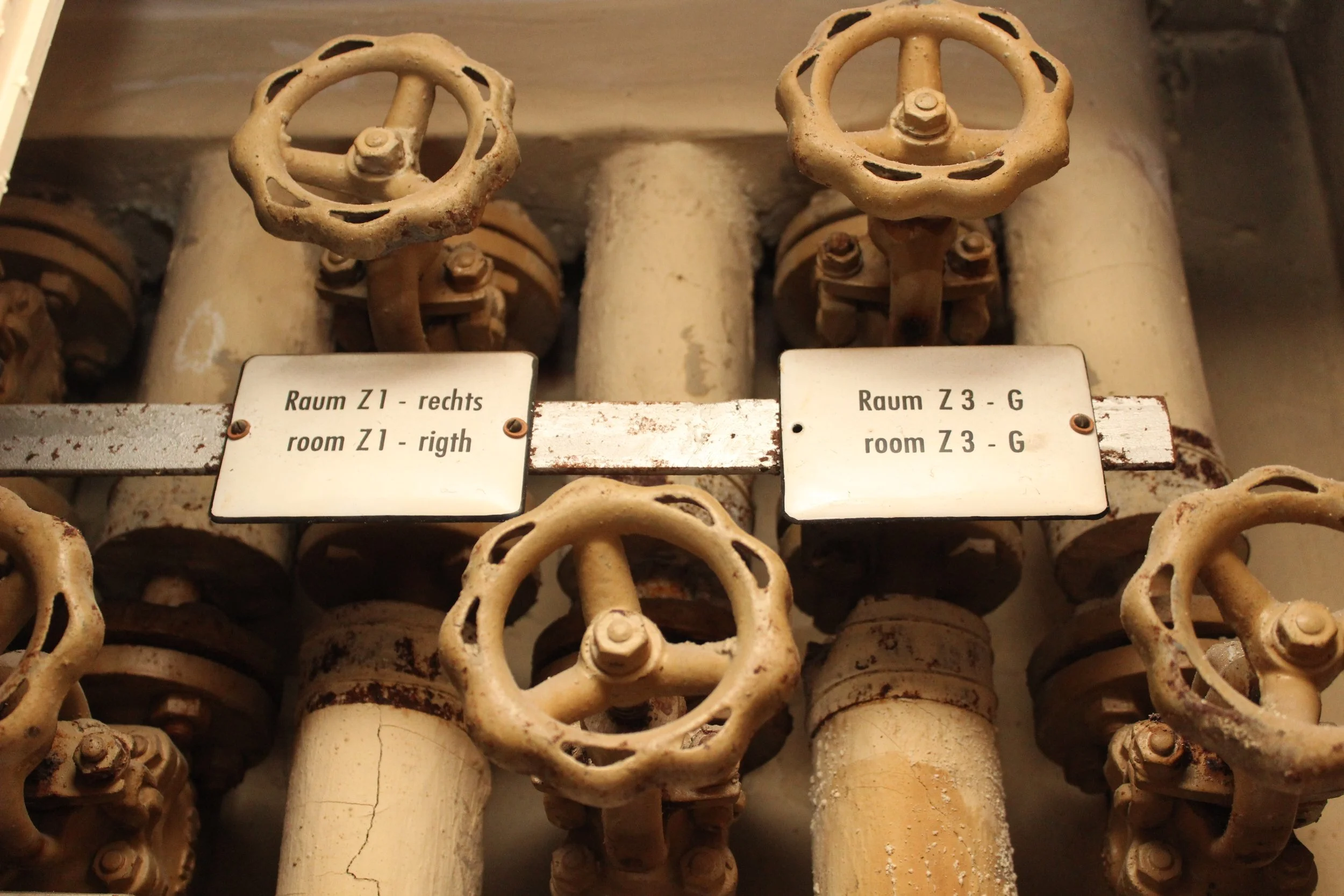 Alte Wasserrohre mit Ventilen und Beschilderung in Deutsch und Englisch, die die Zimmerbezeichnungen und Richtungen anzeigen.