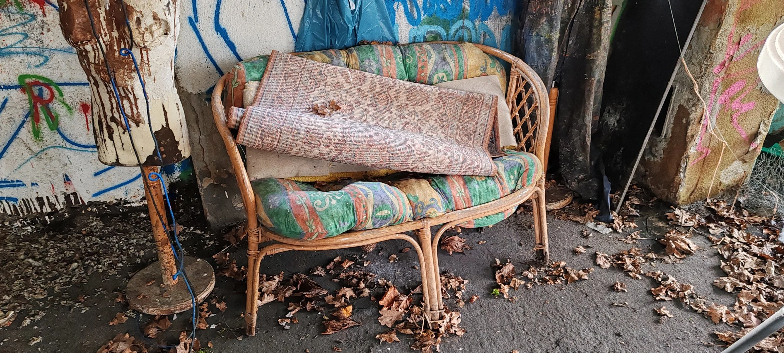 Ein alter Korbsofa mit buntem Polster, das von einem Teppich bedeckt ist, befindet sich im Freien. Der Boden ist mit trockenen Laubblättern bedeckt, und die Wand im Hintergrund ist mit Graffiti bemalt.