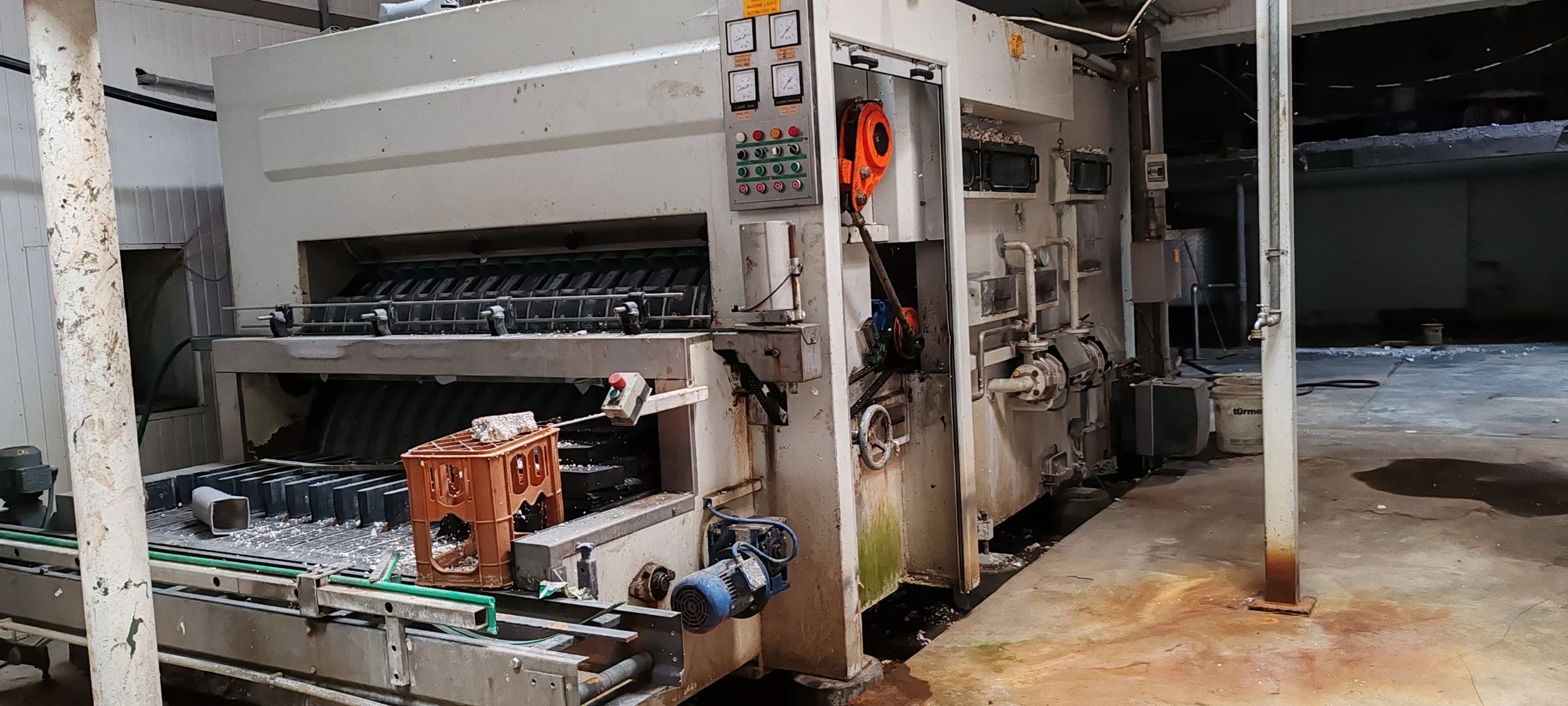 Industrieanlage in einer Fabrik mit Steuerpulten, hölzerner Kiste und Metallrohren, in einem Raum mit schmutzigem Boden.