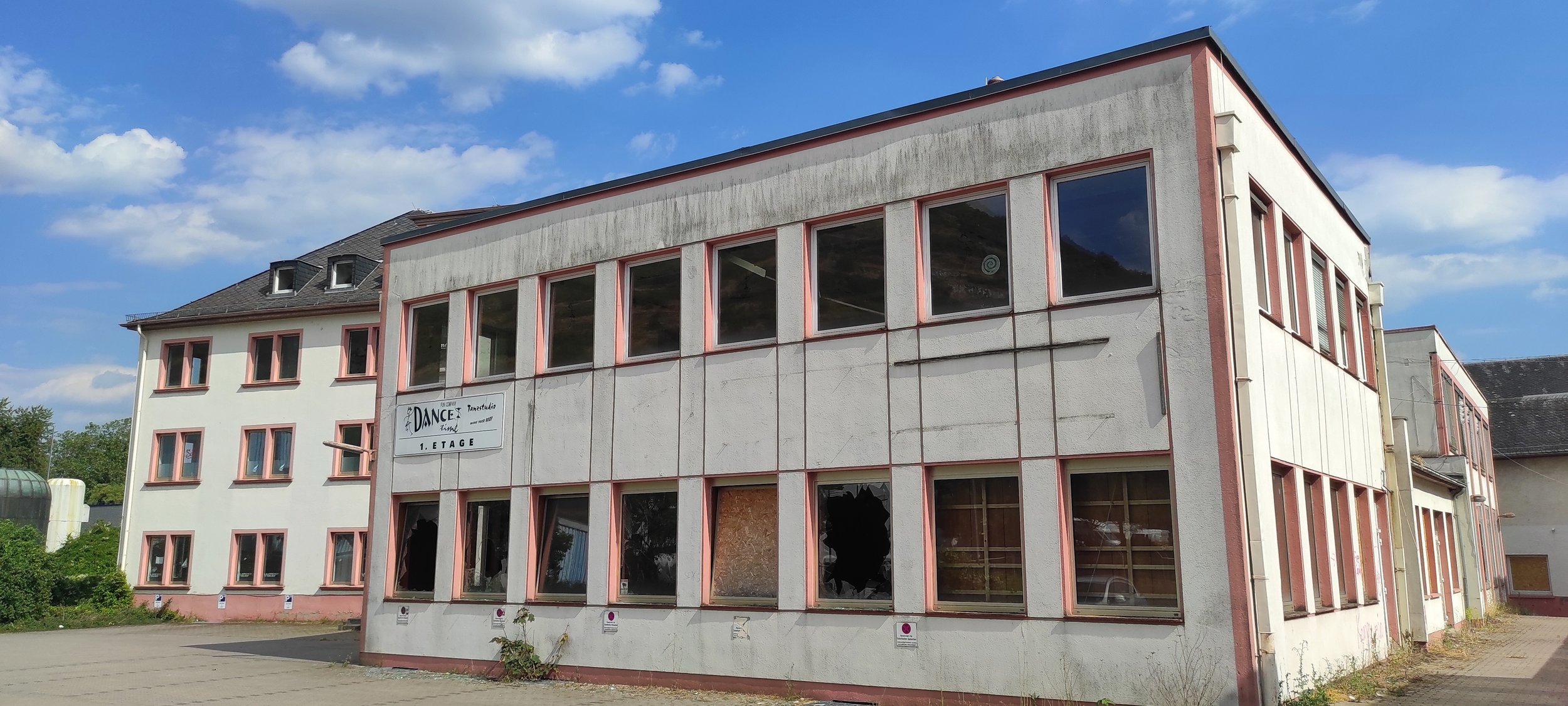 Ein mehrstöckiges Gebäude mit weißen Wänden und rosafarbenen Rahmen um die Fenster, das anscheinend renoviert oder unvollendet ist. Es hat mehrere große Fenster, manche sind zerbrochen oder abgedeckt. Es gibt einen Parkplatz im Vordergrund und einen blauen Himmel mit wenigen Wolken im Hintergrund.