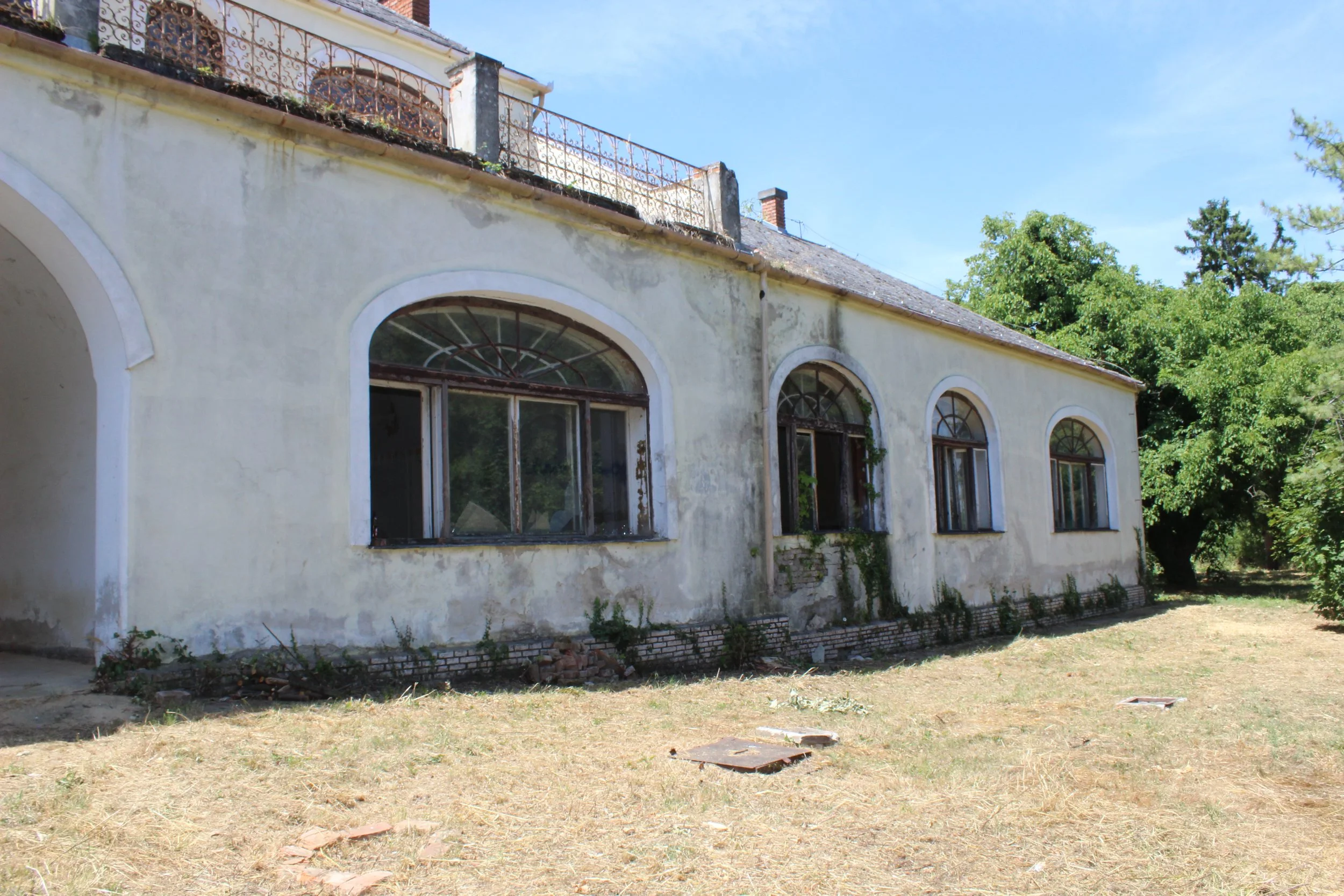 Altes, verfallenes Gebäude mit großen bogenförmigen Fenstern und grünem Bewuchs an den Wänden, umgeben von trockenem, unkrautigem Gras und Bäumen