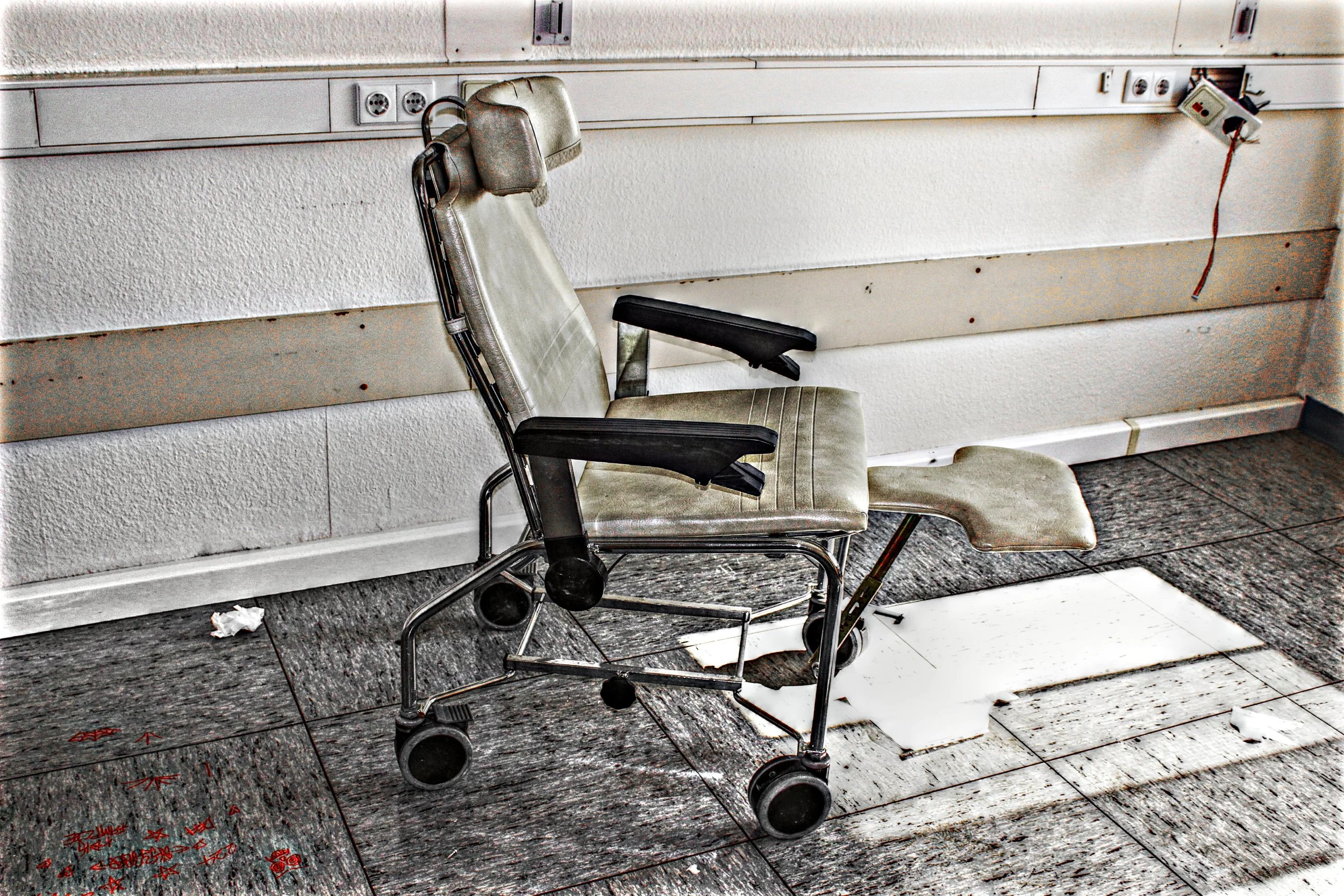 Ein alter, abgenutzter Krankenhaus- oder Rollstuhl im verlassenen, unordentlichen Raum mit beschädigten Fliesen am Boden.