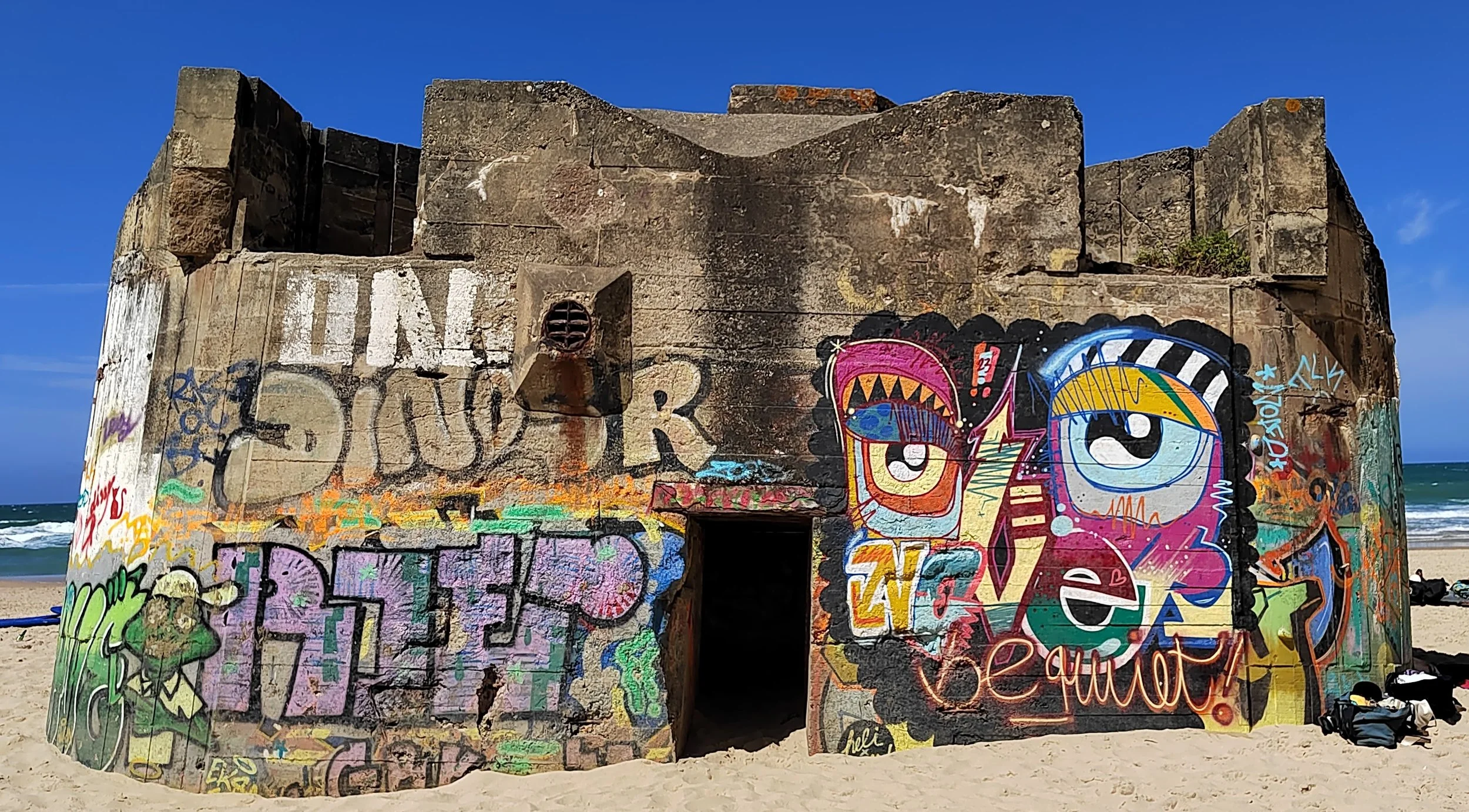 Verfallene Bunker mit buntem Graffiti am Strand, im Hintergrund das Meer und blauer Himmel.