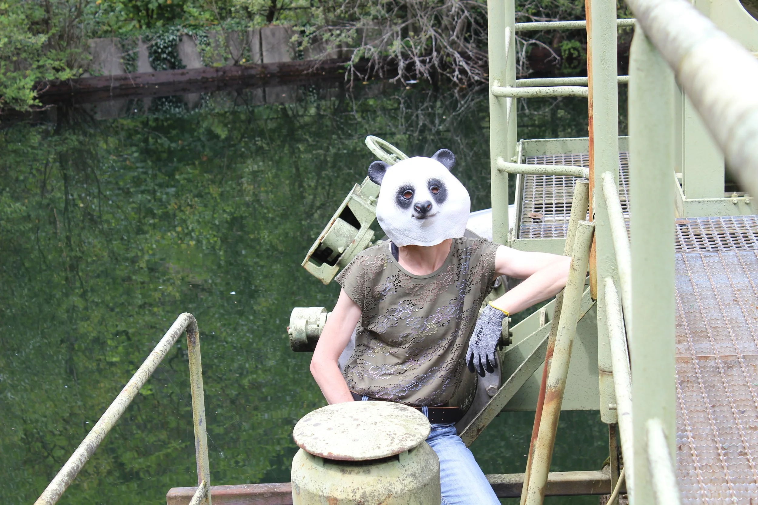 Person mit Affenkopf-Maske auf einer alten, grünen Metalltreppe an einem Fluss, umgeben von Bäumen.
