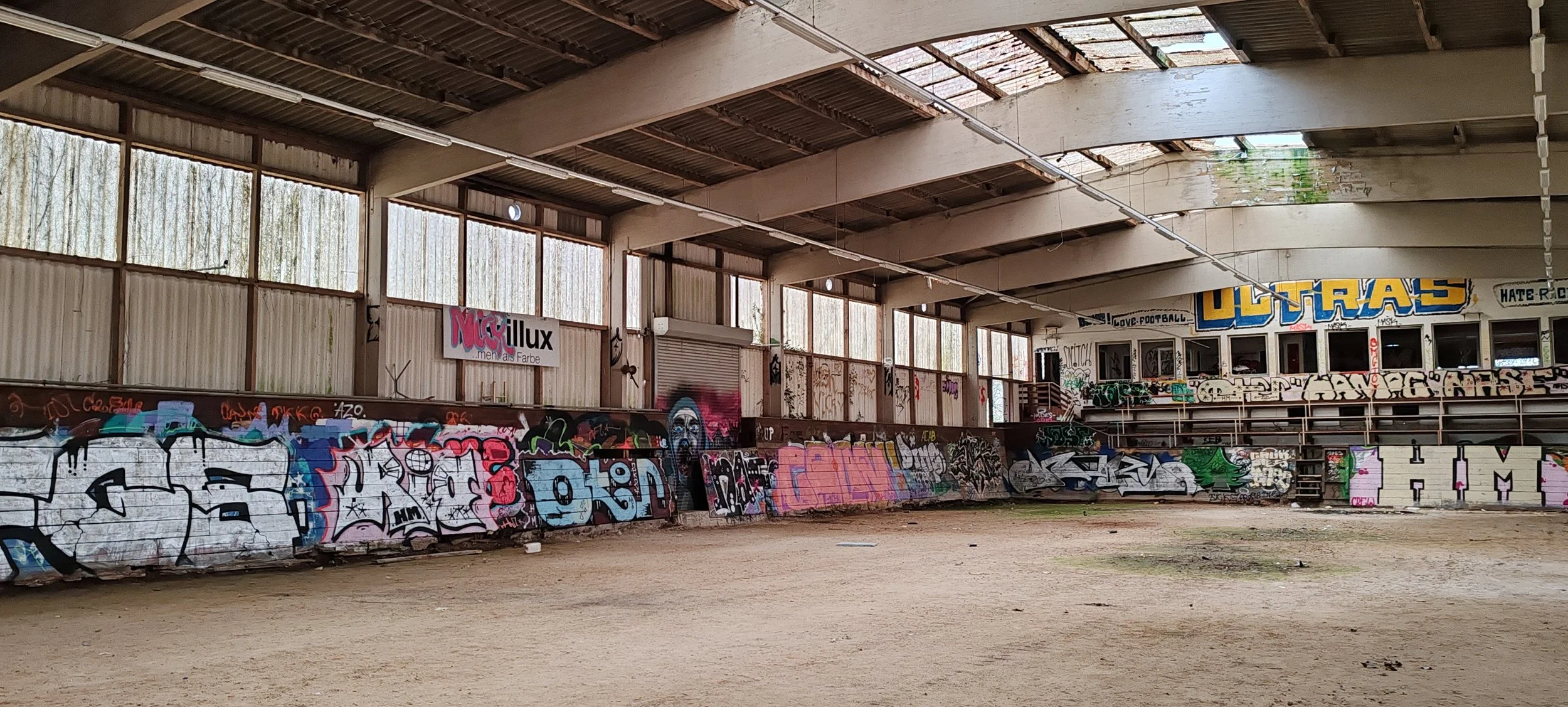 Verlassener Indoor-Sportplatz mit Graffiti auf den Wänden und in mehreren Fenstern. Der Boden ist staubig und uneben.