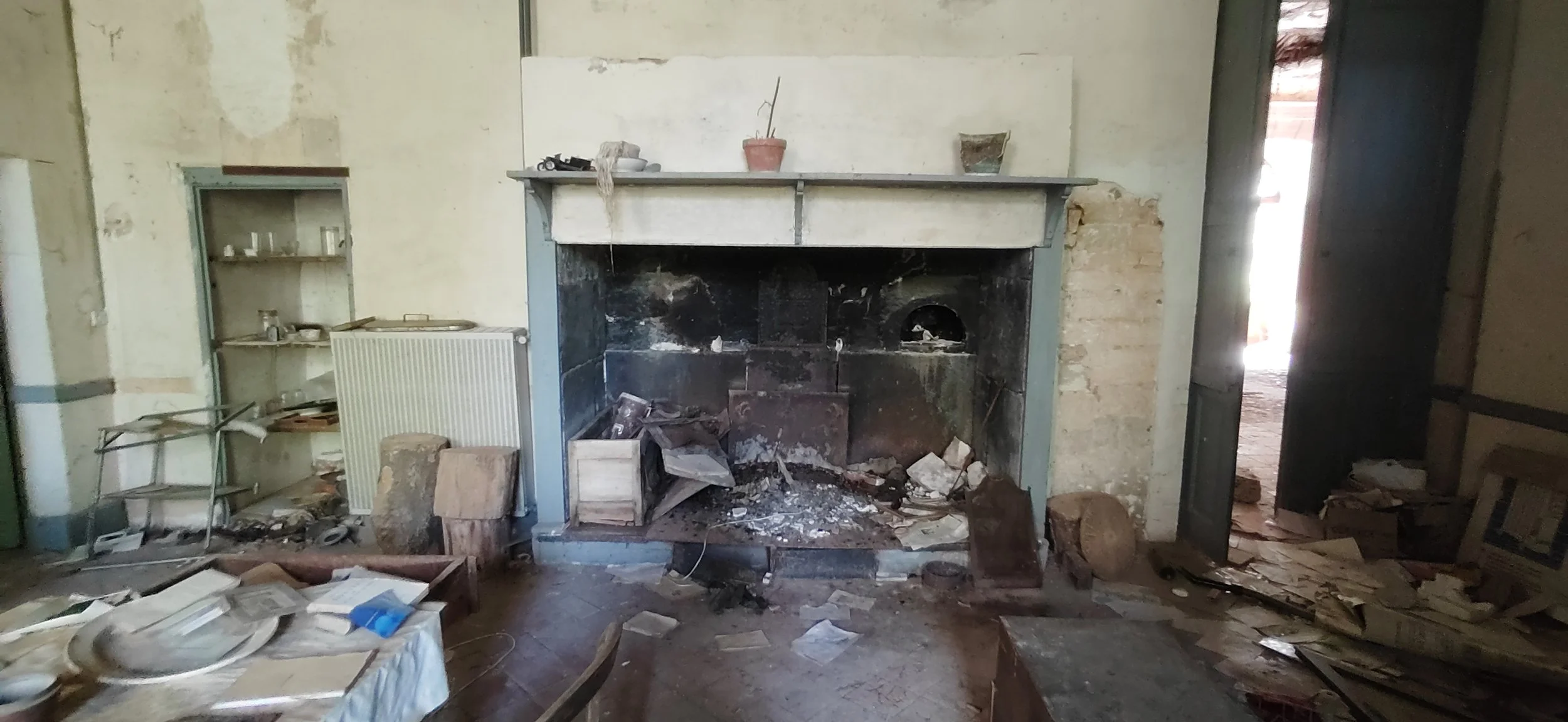 Ein alter, abgenutzter Kamin in einem verwohnten Raum, umgeben von Schmutz, zerbrochenem Holz, Staub und Terrakotta-Töpfen auf dem Kaminsims.