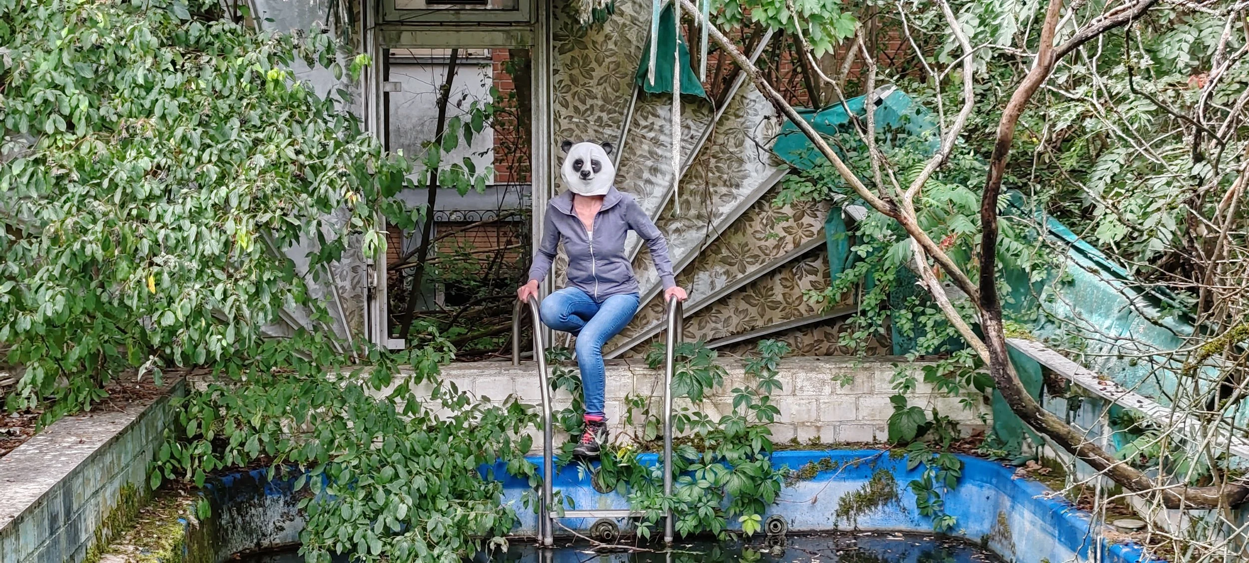 Eine Person mit einer Panda-Maske, die in einer grauen Jacke und blauen Jeans sitzt, auf einer Metallleiter vor einer verfallenen Gartenpoolszene mit Pflanzen und Dschungelvegetation.