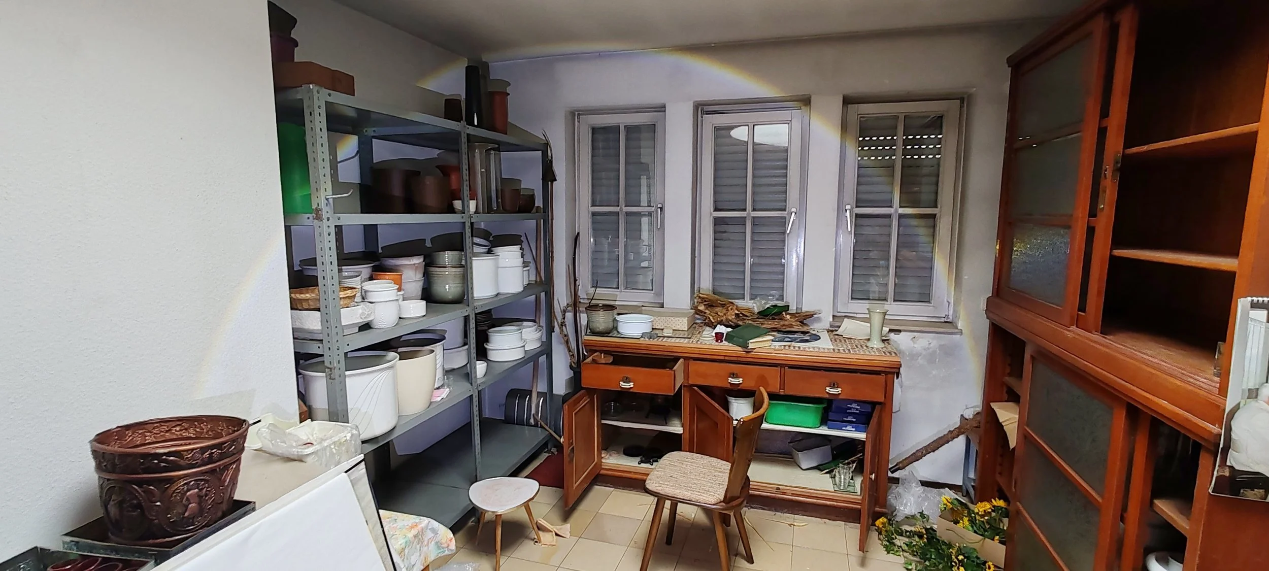 Ein Raum mit Geschirr, Holzmöbeln, einer Wetterkappe, mehreren Fenstern, verschiedenen Pflanzen und Gegenständen auf den Möbeln, die Umgebung wirkt unordentlich.