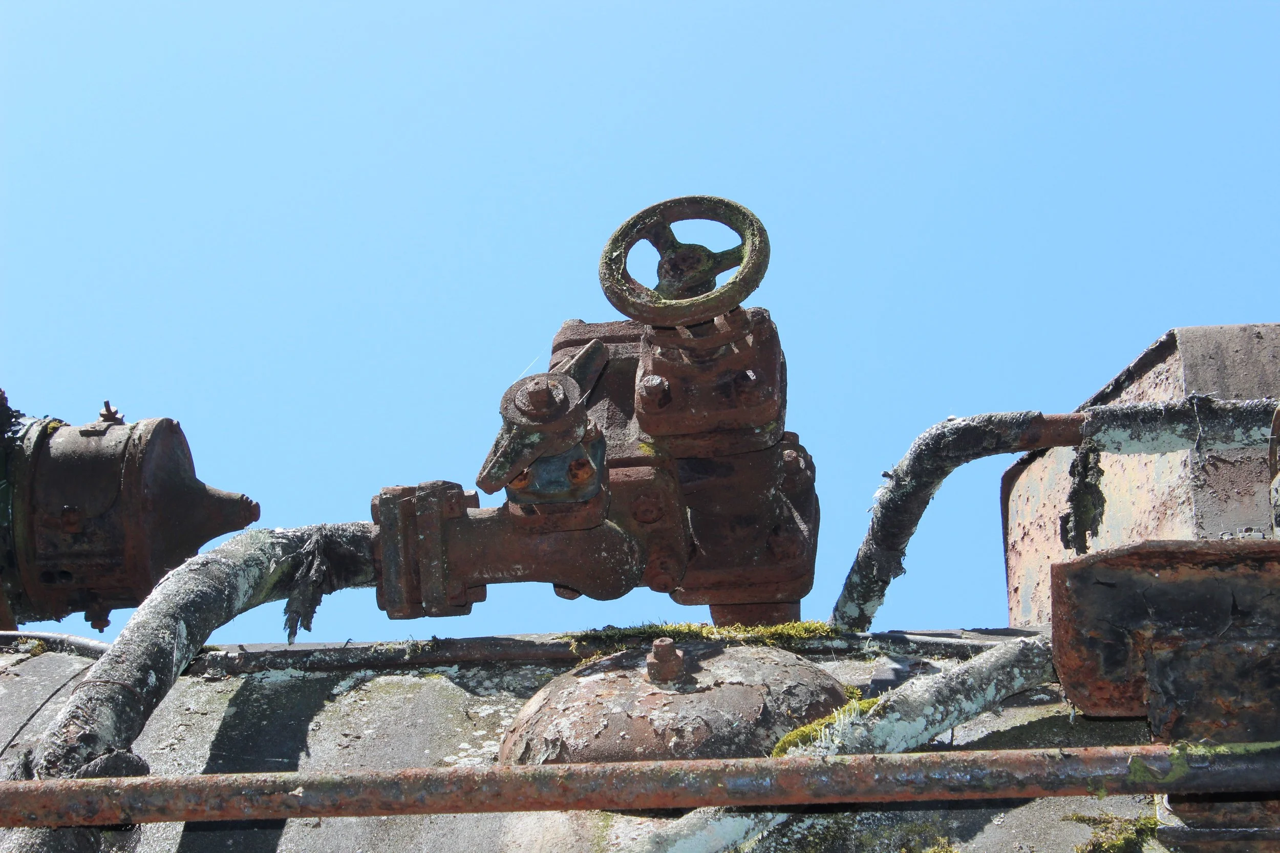 Rostige industrielle Rohrleitungen und Ventile auf einem alten Dach, mit deutlich sichtbaren Rost und Moos, gegen einen klaren blauen Himmel.