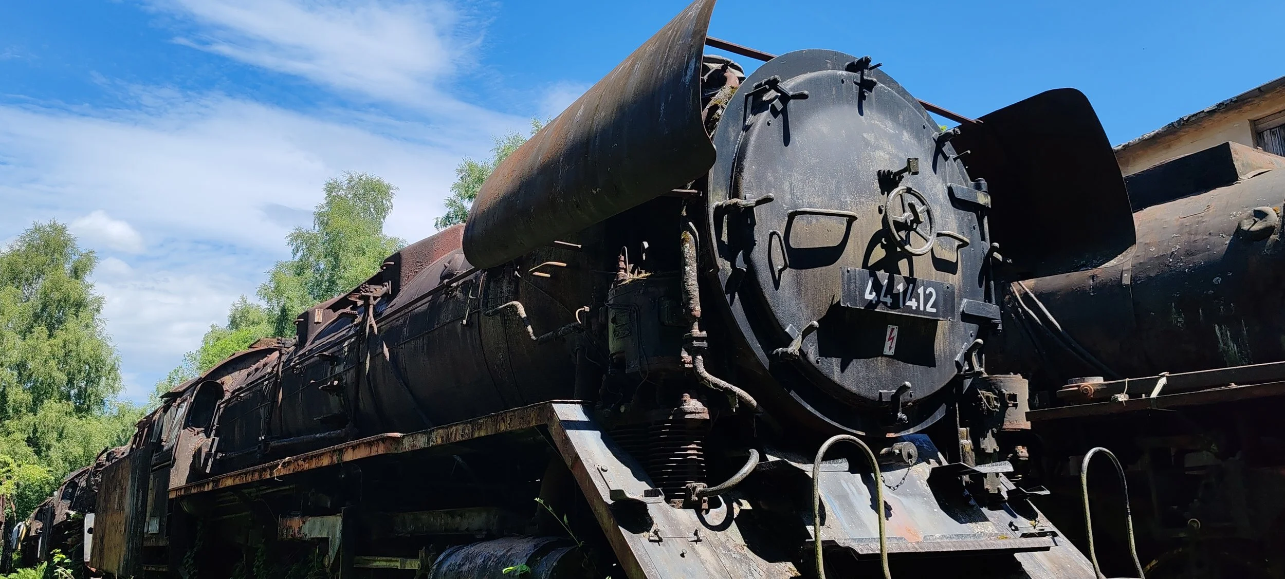 Verrostete alte Dampflokomotive im Freien, umgeben von grünen Bäumen und blauen Himmel.