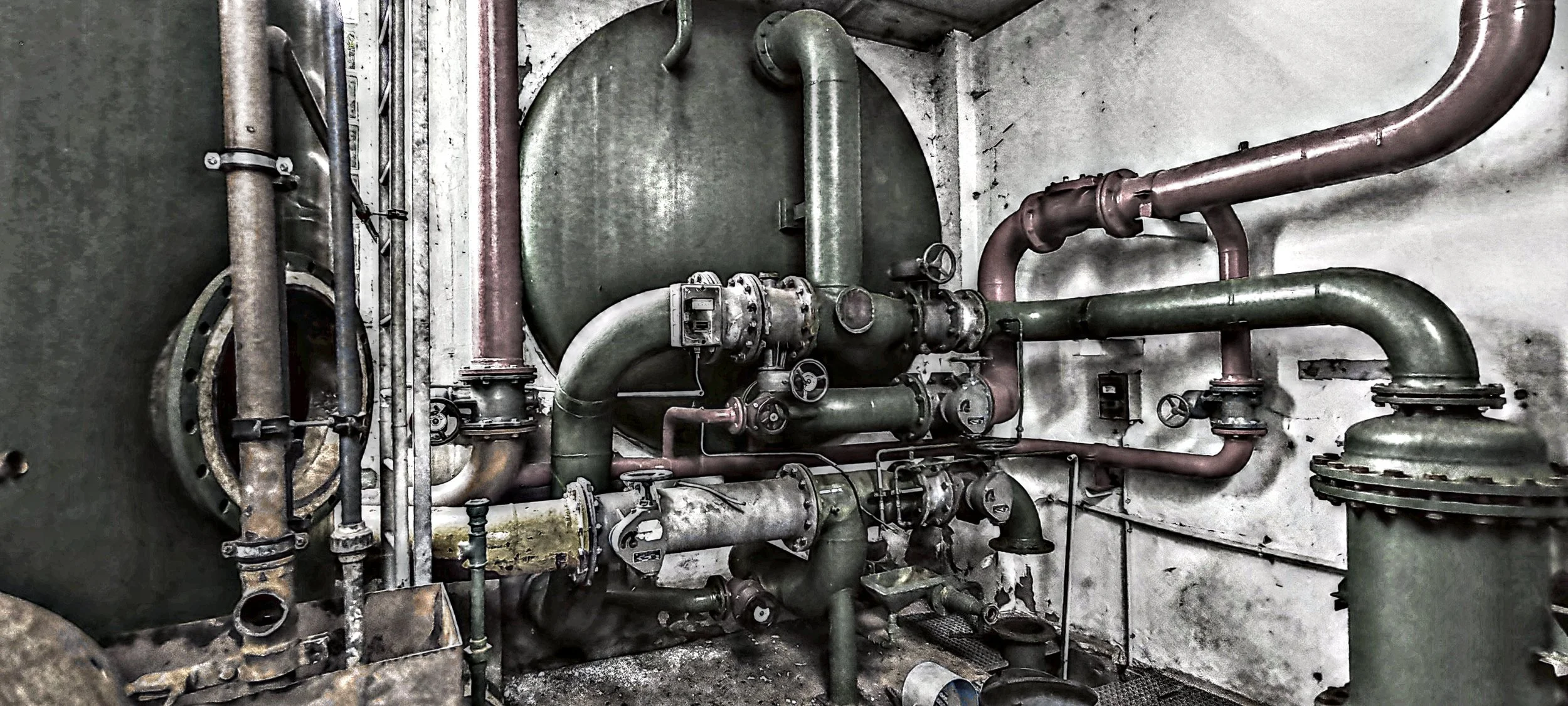 Rostfreie, alte Industriemaschinen und Rohrleitungen in einer dunklen, abgenutzten Fabrikhalle.