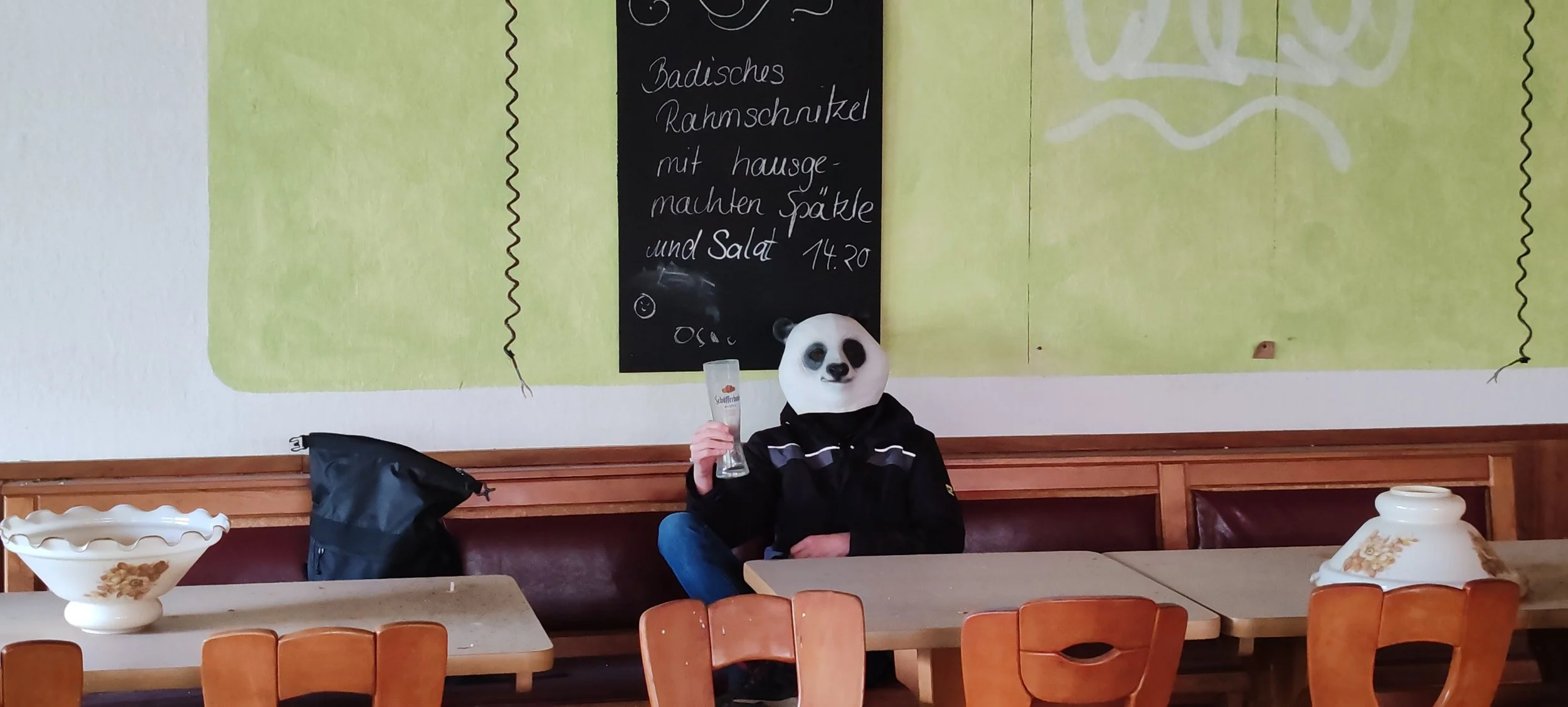 Eine Person mit Panda-Maske sitzt an einem Tisch in einem Restaurant oder Café, hält ein Glas und lehnt sich an die Wand. Es gibt zwei große Keramikvasen auf den Tischen und eine Tafel mit handgeschriebenem Menü im Hintergrund.