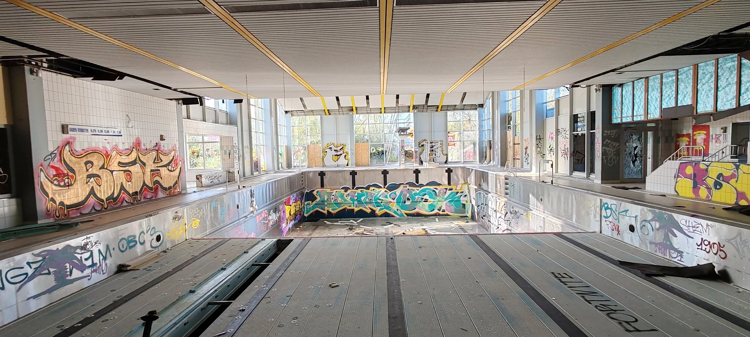 Ein leerer, verfallener Badebereich mit Graffiti an den Wänden und an der Beckenwand, große Fenster lassen natürliches Licht herein, und das Wasser im Becken ist abgestanden.
