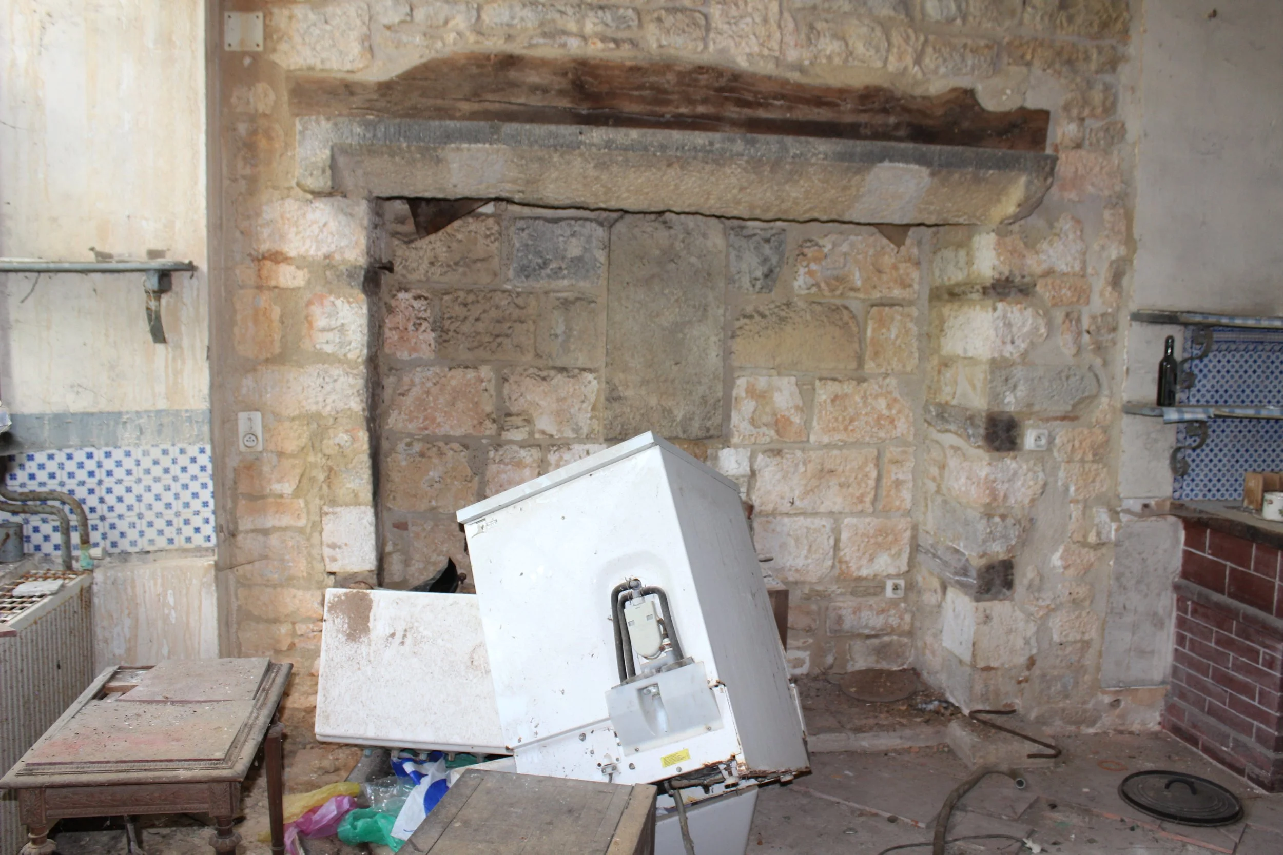 Innenraum einer alten, teils renovierten Küche mit beschädigtem weißen Kühlschrank, umgeben von Baustellenmaterialien und Werkzeugen.