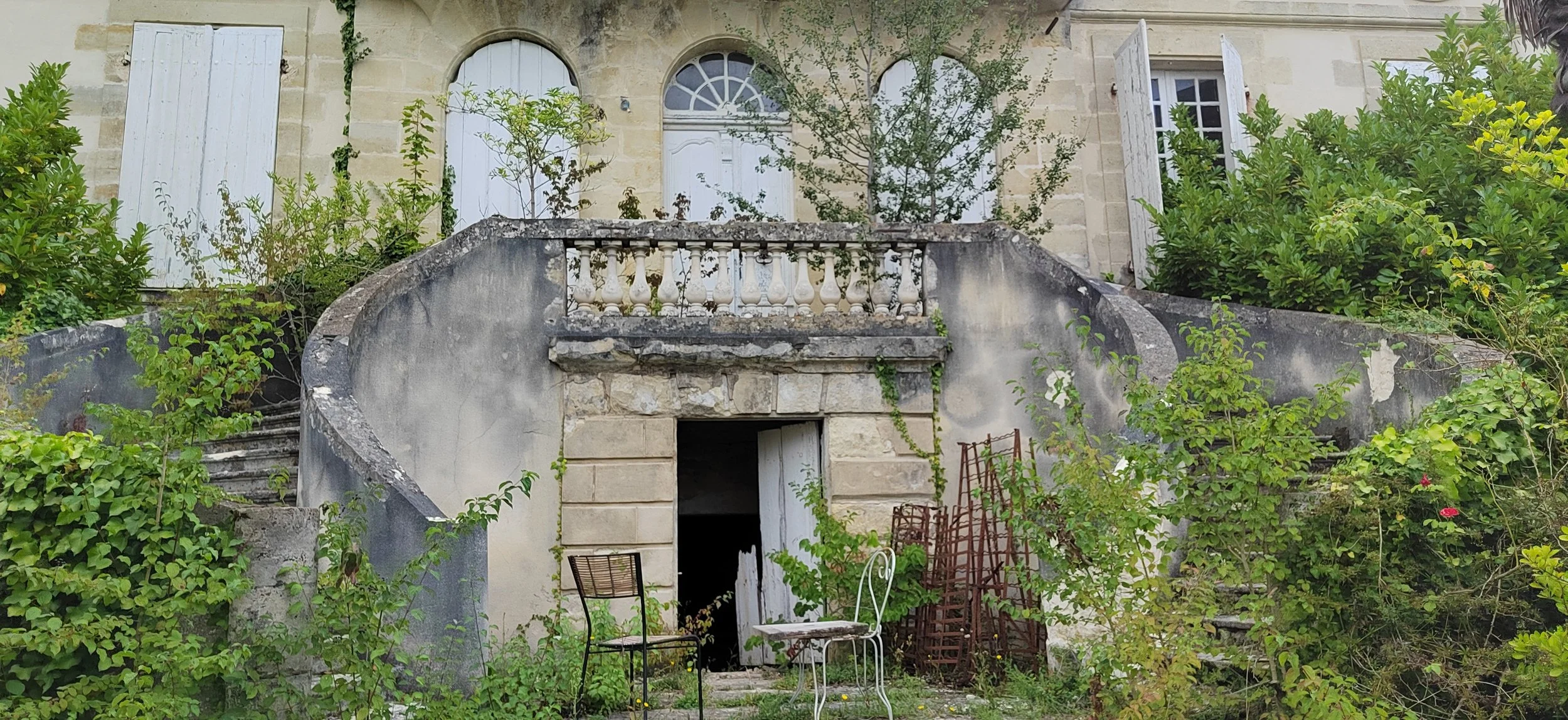 Verfallene Villa mit offener Tür, umgeben von Pflanzen, Stühlen und Gartenmöbeln im Garten.