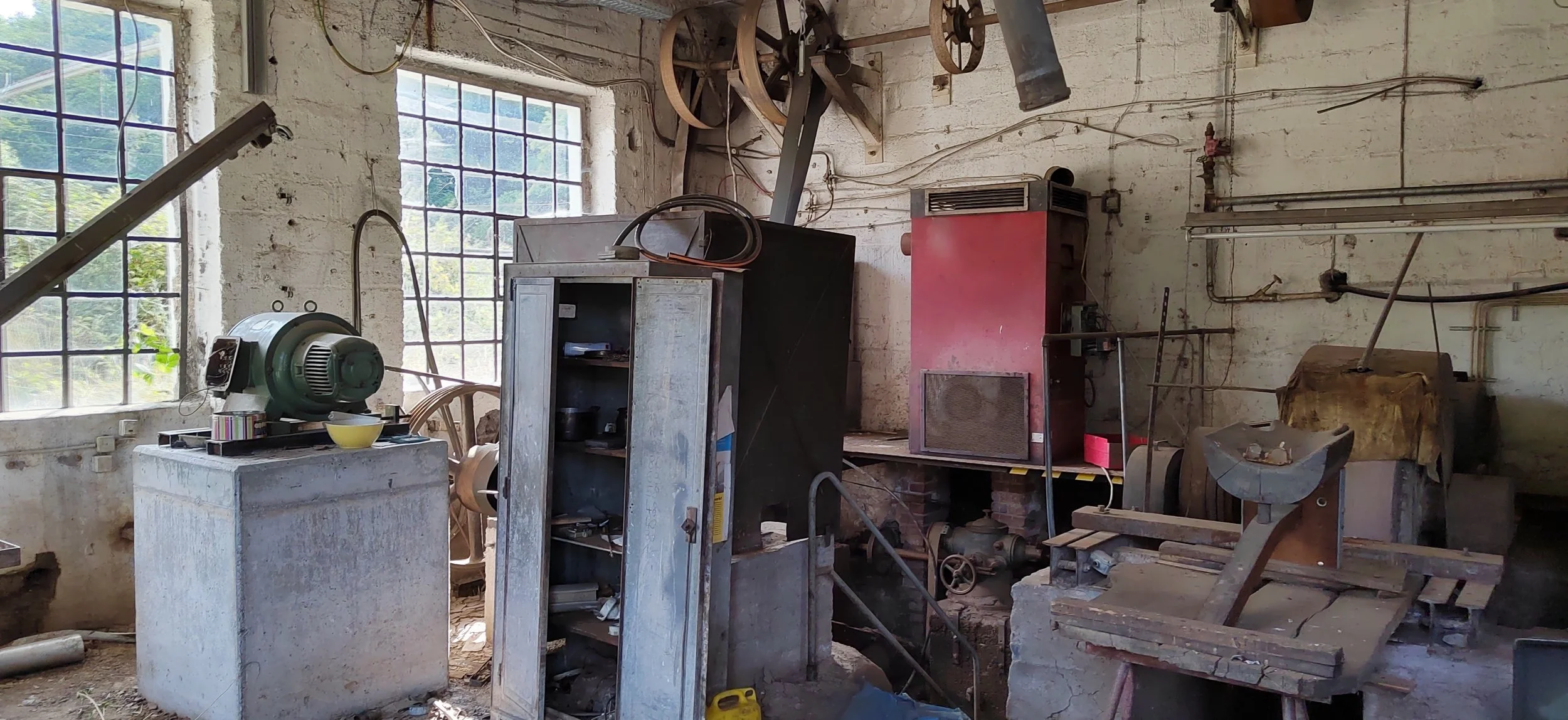 Ein alter, verlassener Werkstattraum mit verschiedenen Maschinen, Werkzeugen und Lagerregalen, die teilweise verrostet und schmutzig sind.