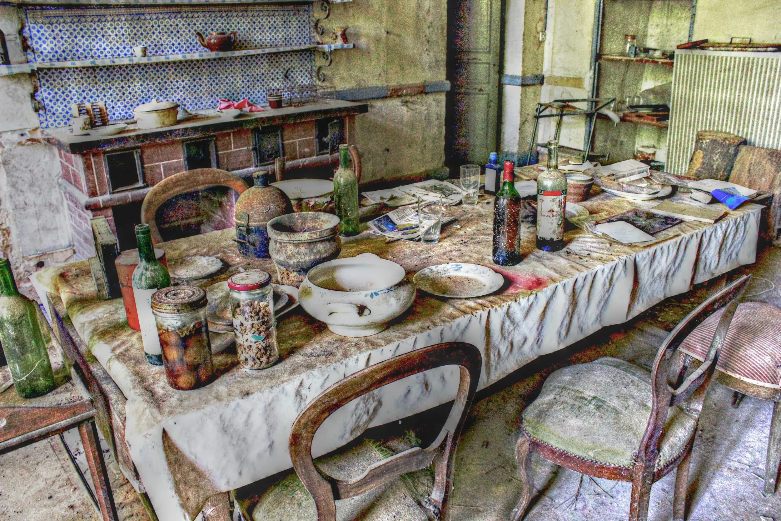 Ein verwaister Esstisch mit schmutzigem Tischtuch, zerbrochenen und verrosteten Geschirrresten, leeren Weinflaschen, Keramikvasen und Staub in einer alten, verlassenen Raumkulisse.