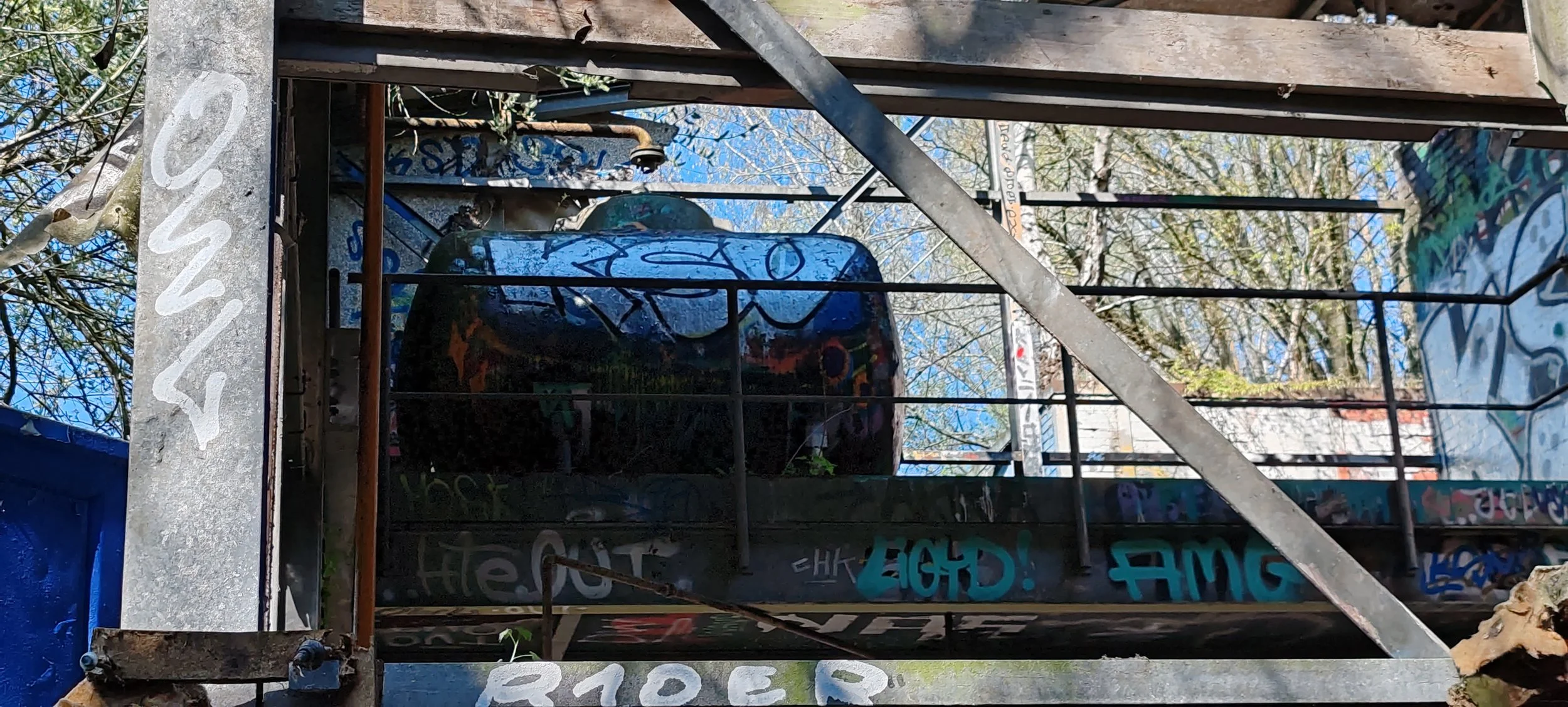 Ein verfallener, graffitiüberdeckter Truck im Freien, umgeben von Bäumen und Metallstrukturen.