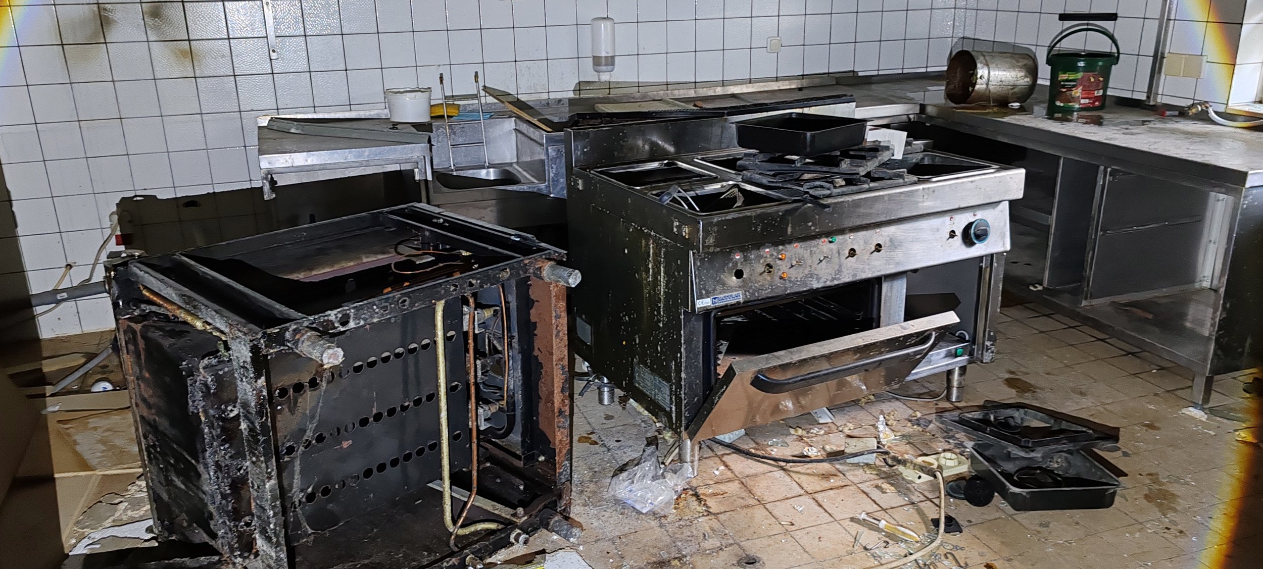 Ein stark beschädigter und verschmutzter Gastronomiebereich mit verstuemmelten, verbrannten und zerbrochenen Kochgeräten, sowie verschmutzter Arbeitsfläche und Boden.