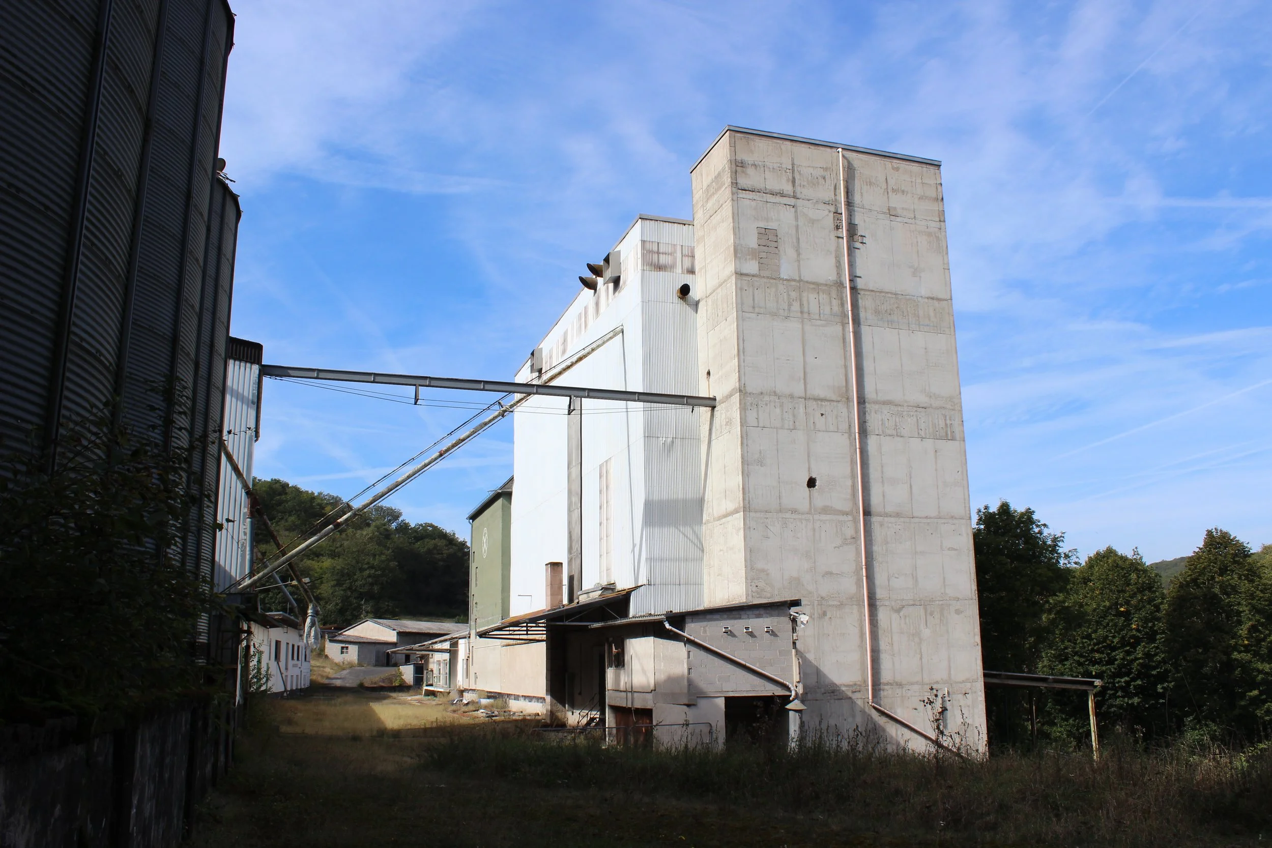 Ein Industriebau mit großen Betonwänden, Rohren und Staubfässern, umgeben von Bäumen und einer unbefestigten Fläche, bei blauem Himmel.
