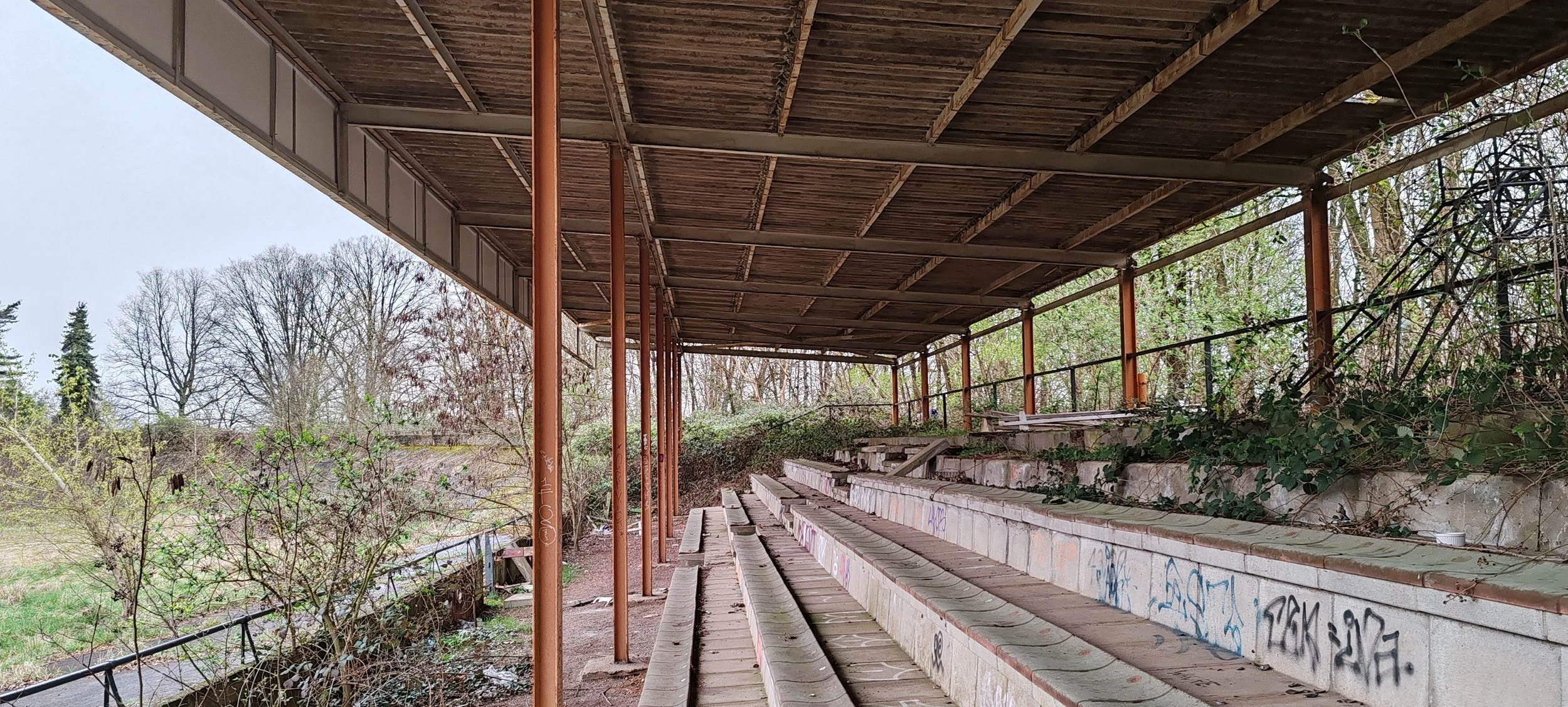 Verfallene, verstaubte Sitzreihen in einer alten, verlassenen Sporttribüne im Freien, umgeben von Bäumen und Graffiti