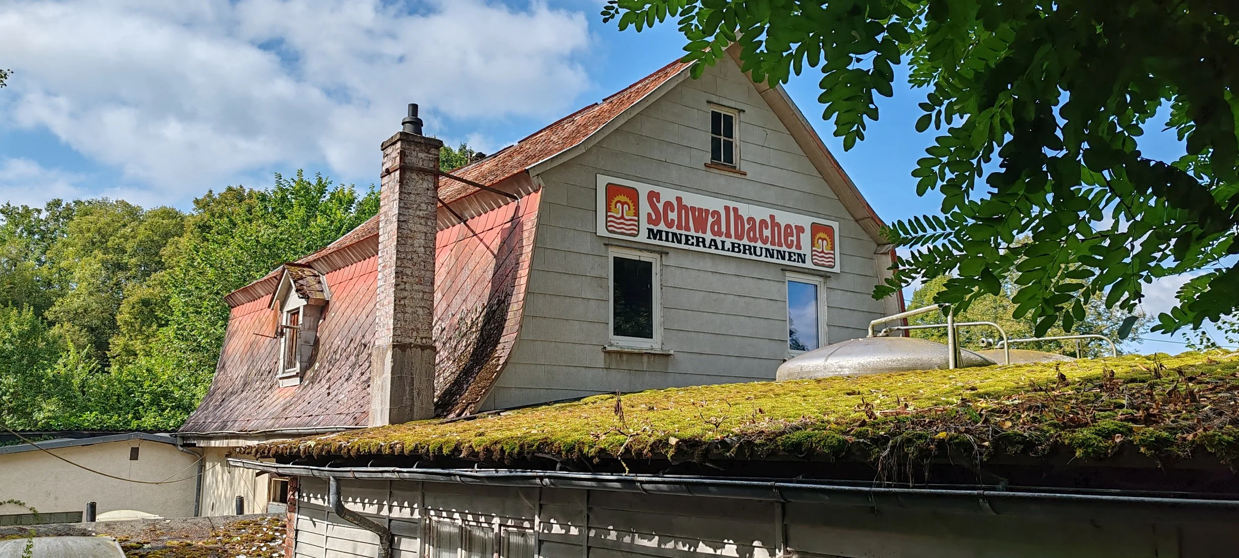 Ein Haus mit einem Schild "Schwalbacher Mineralbrunnen" an der Fassade, umgeben von grünen Bäumen, mit einem verschmutzten, mossbedeckten Dach und einer blauen Himmel mit Wolken.