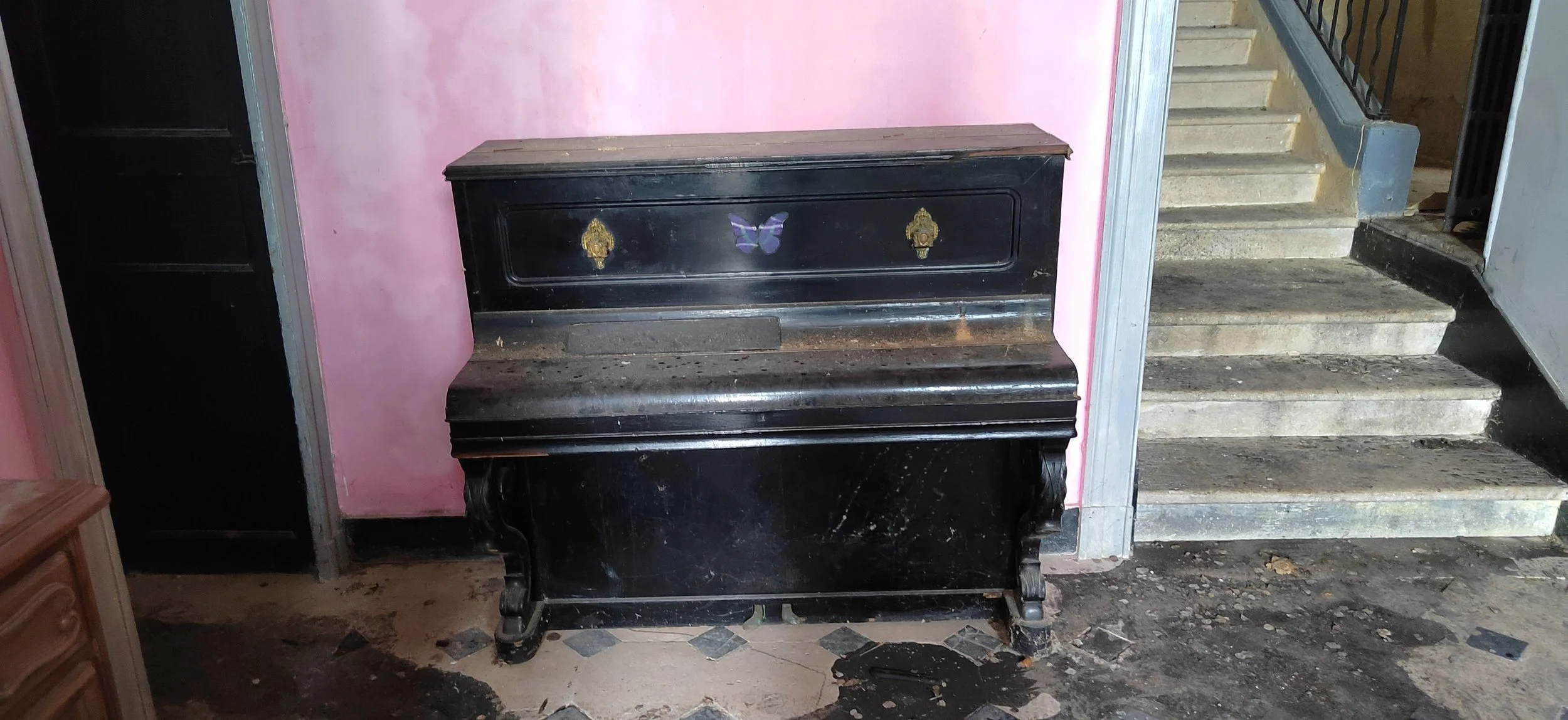 Ein alter, schwarzer Flügel mit Verzierungen, darunter ein rosa Wand, ein Treppenabsatz und eine steinerne Treppe.