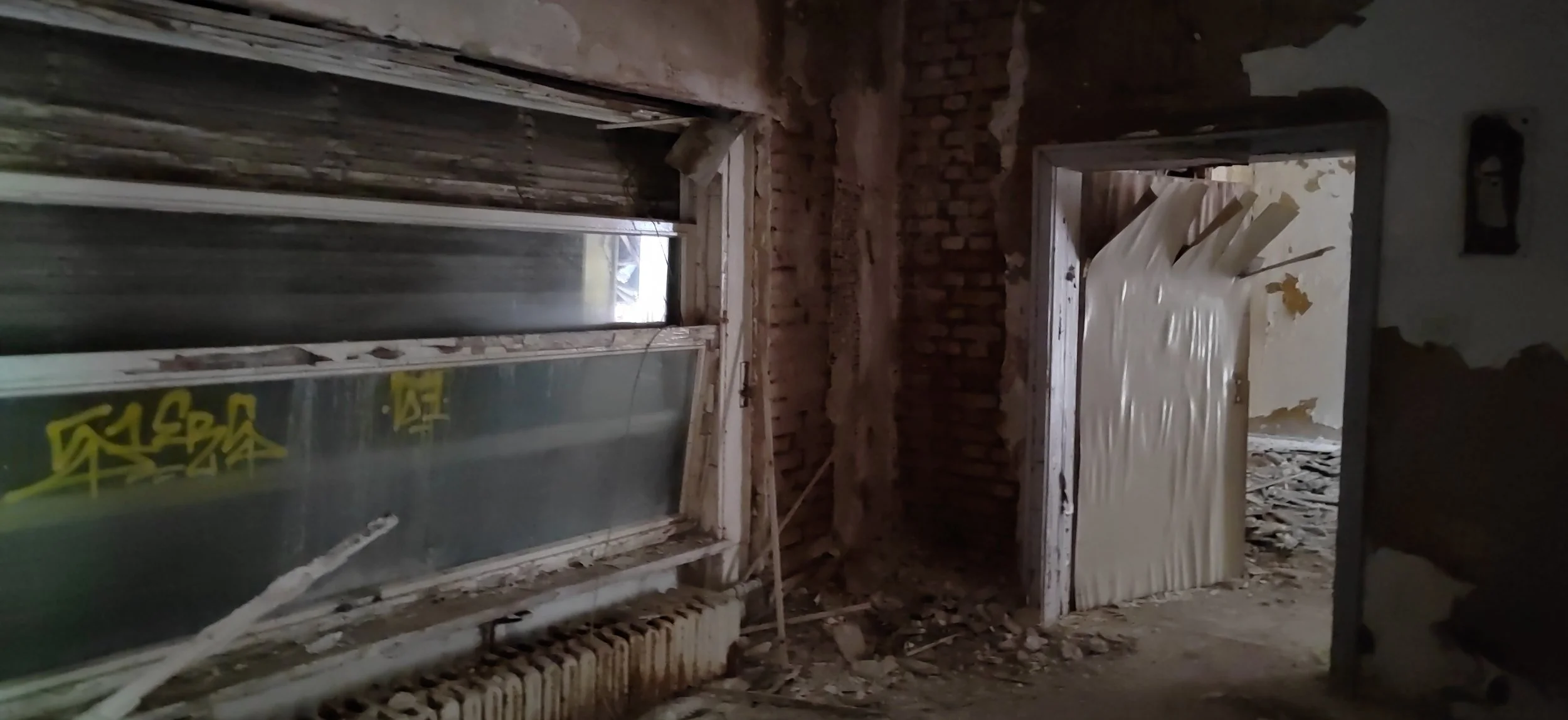 Innenansicht eines Baustellenraums mit einem Fensterrahmen, einer geöffneten Tür, die mit Kunststofffolie abgedeckt ist, und ungeordneter Bauschutt auf dem Boden.