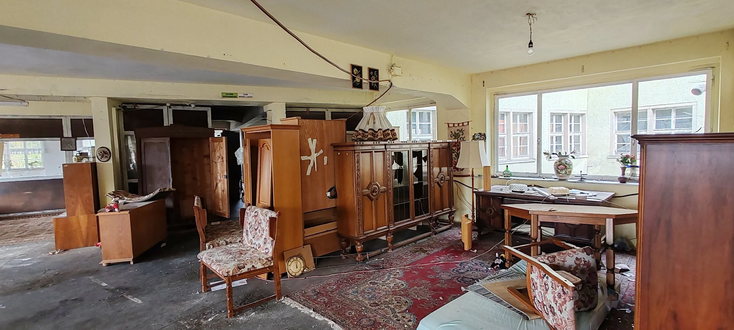 Ein chaotischer Raum mit alten Möbeln, Stühlen, Schränken und einem Teppich vor einer großen Fensterfront.