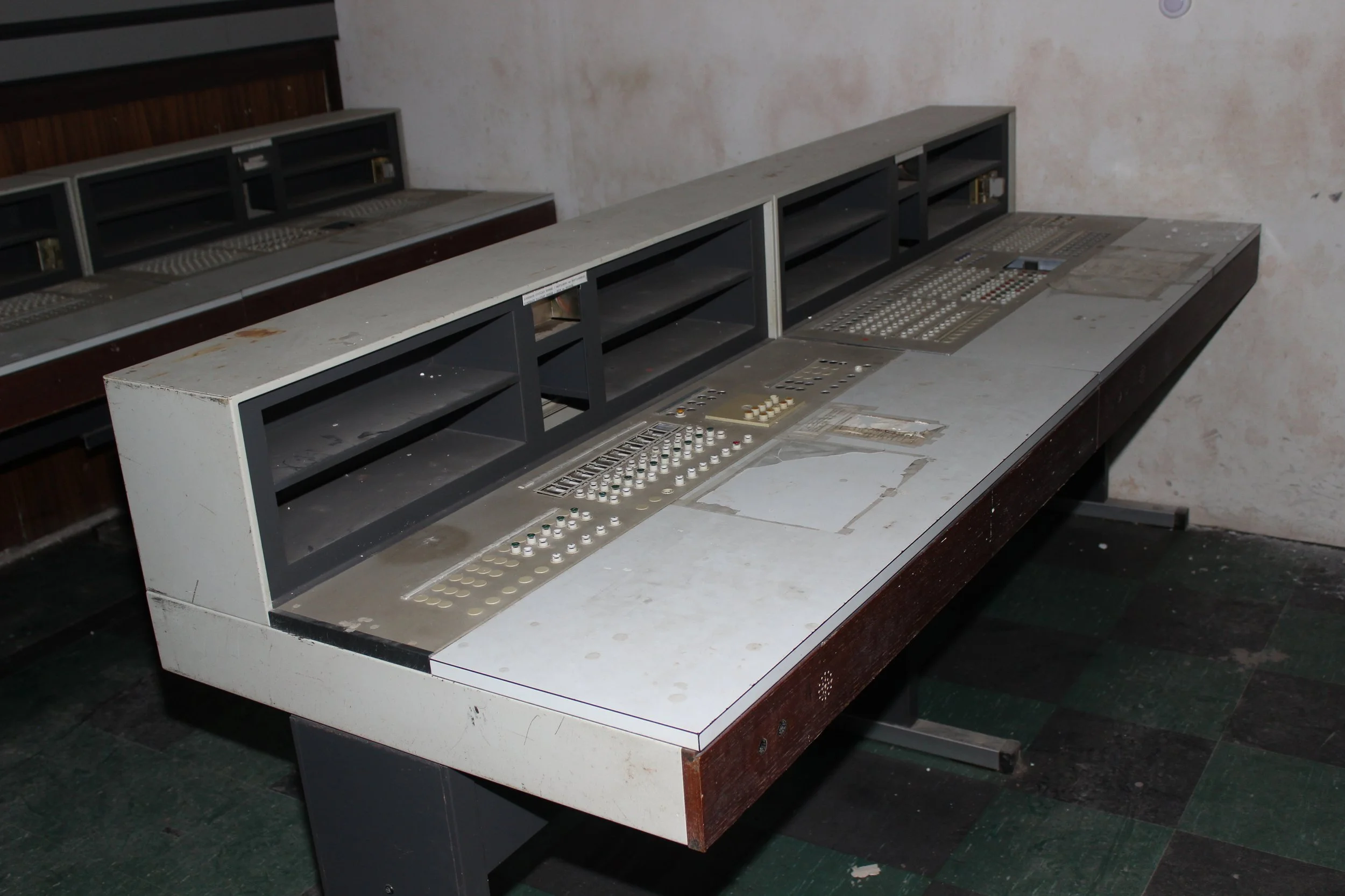 Verlassener und veralteter Kontrollraum mit leeren und verschmutzten Schreibtischen, alten Elektronikgeräten und einer ungeordneten Umgebung.