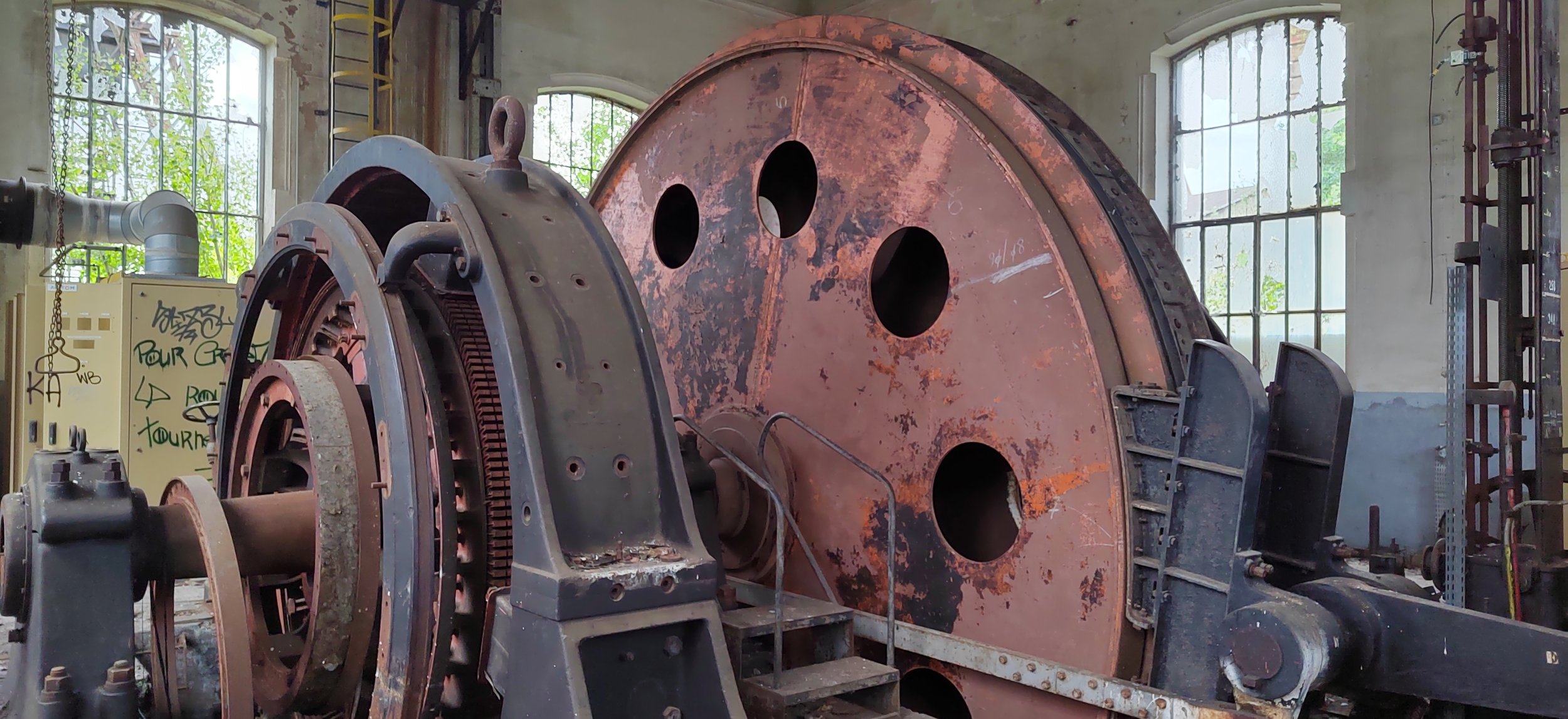 Alter, rostiger Maschinenteil in einer Industriehalle mit großen Fenstern im Hintergrund.